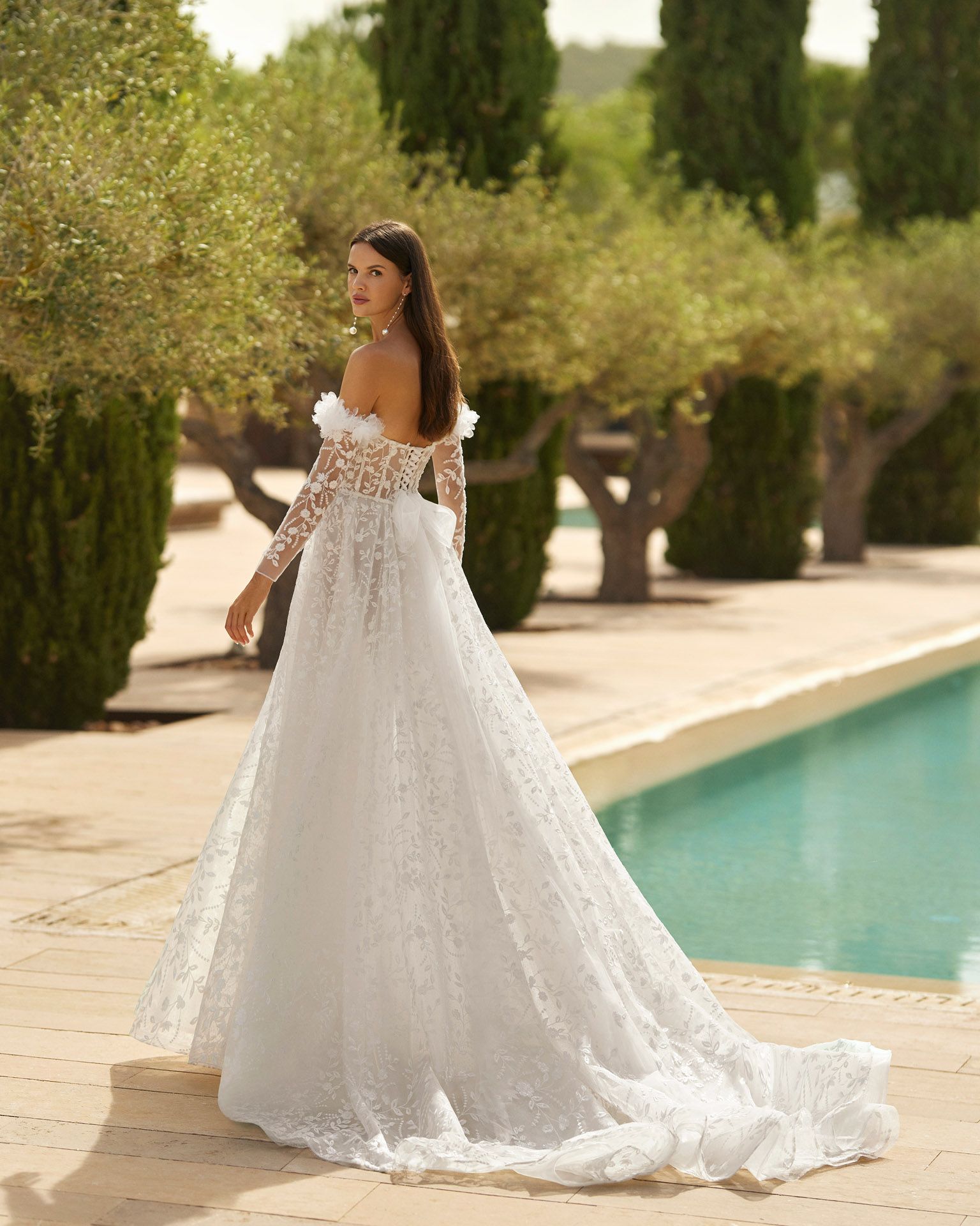 Donna in abito da sposa bianco con lungo strascico, in piedi vicino a una piscina, all'aperto.