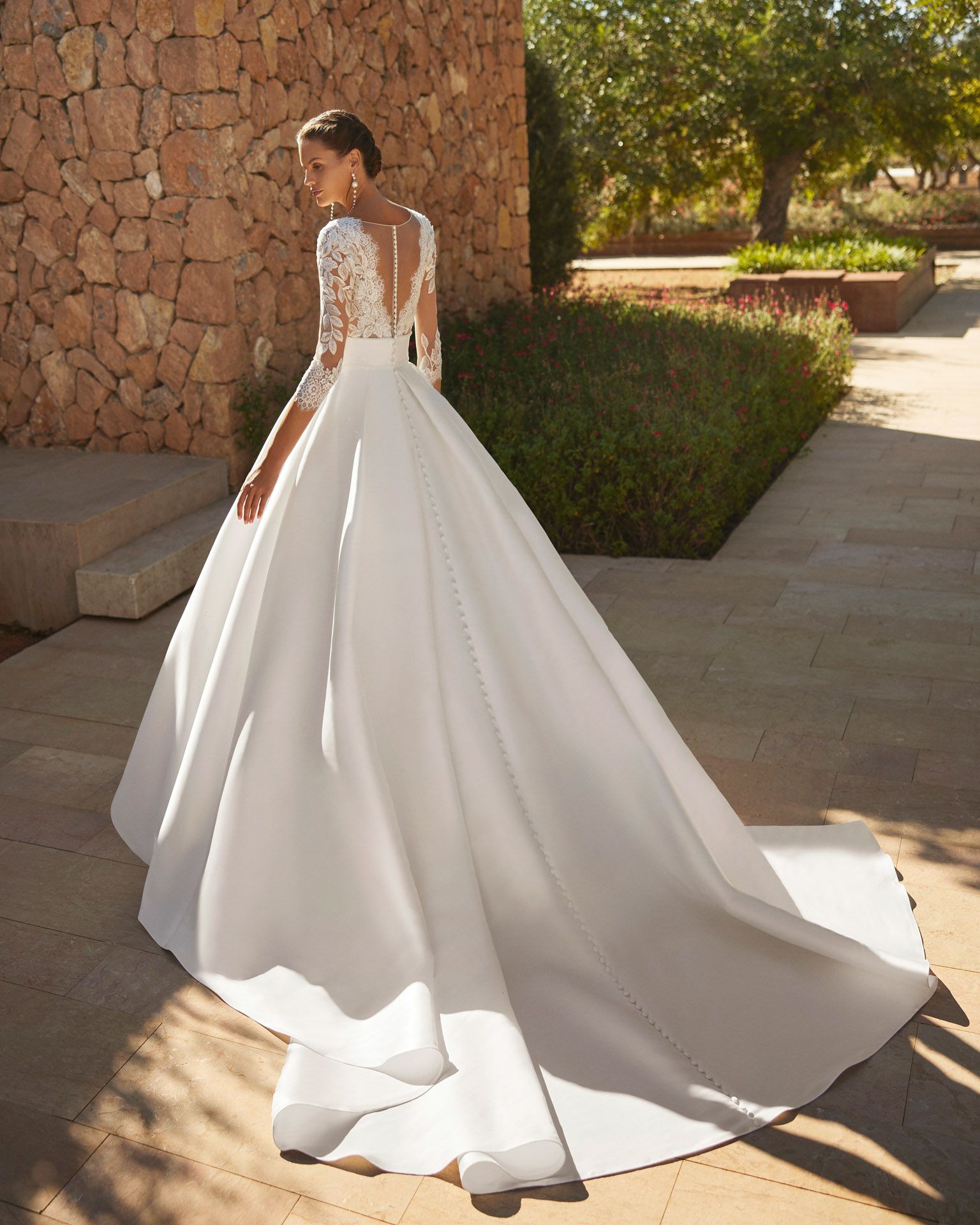 Donna in abito da sposa bianco con lungo strascico, in piedi all'esterno, vicino a un muro di pietra.