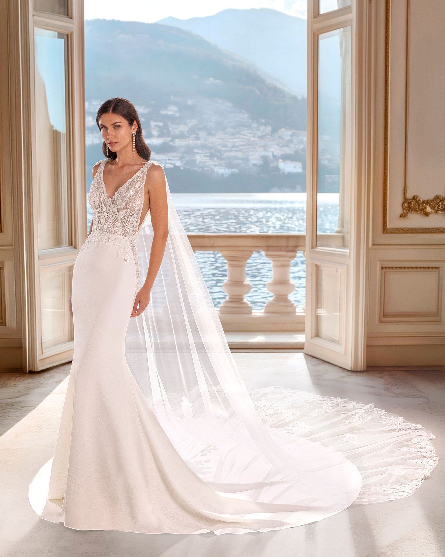 Donna in abito da sposa bianco a sirena con corpetto in pizzo e strascico fluente, in piedi vicino a un balcone con vista sull'acqua e sulle montagne.
