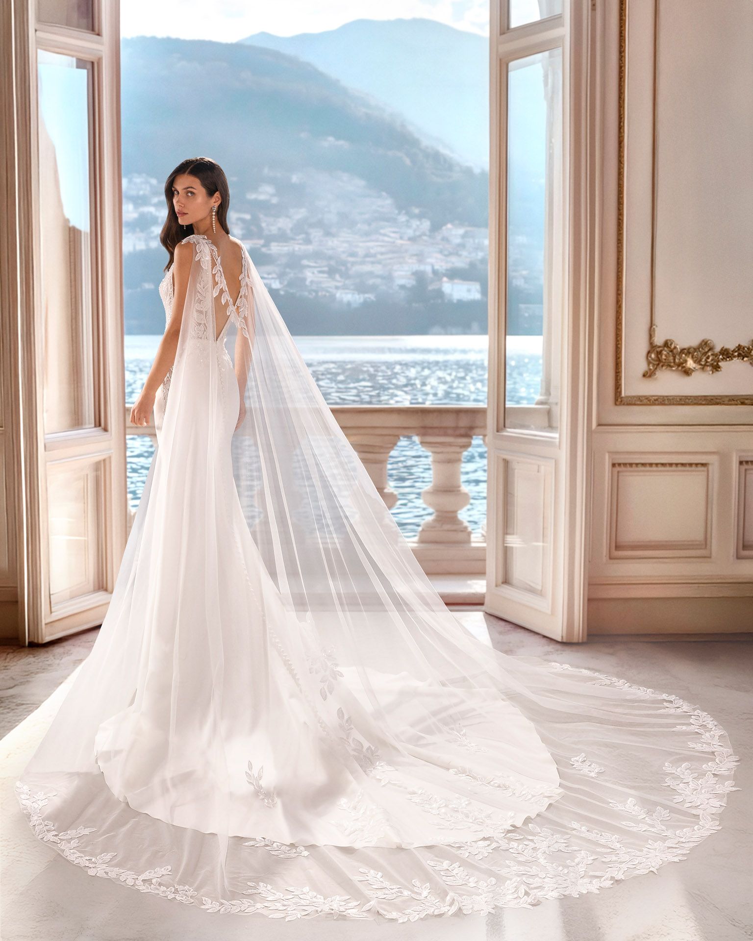 Una donna con un abito da sposa bianco con un lungo strascico è in piedi su un balcone con vista su un lago e sulle montagne.