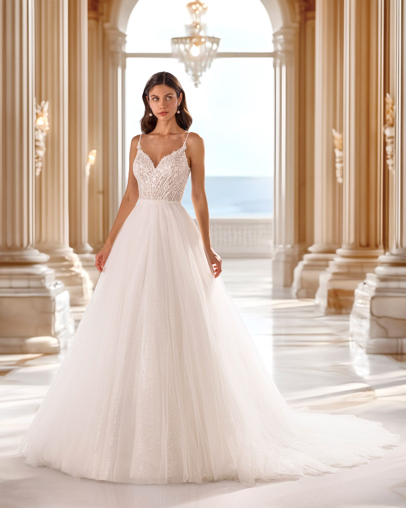 Donna in abito da sposa bianco con spalline sottili, in piedi in una grande sala, che guarda avanti.