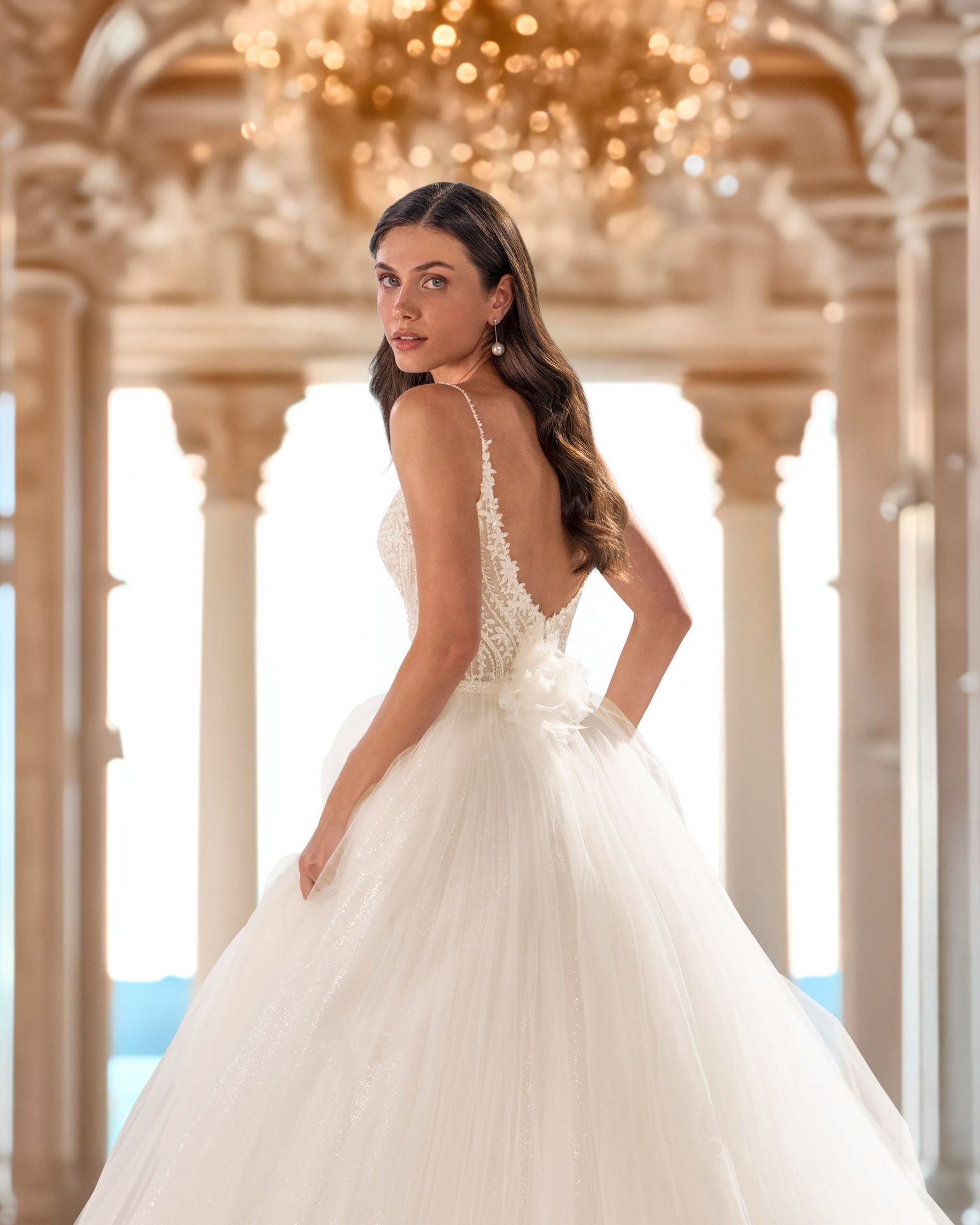 Donna in abito da sposa bianco, che guarda oltre le spalle, in una grande sala con colonne e un lampadario.