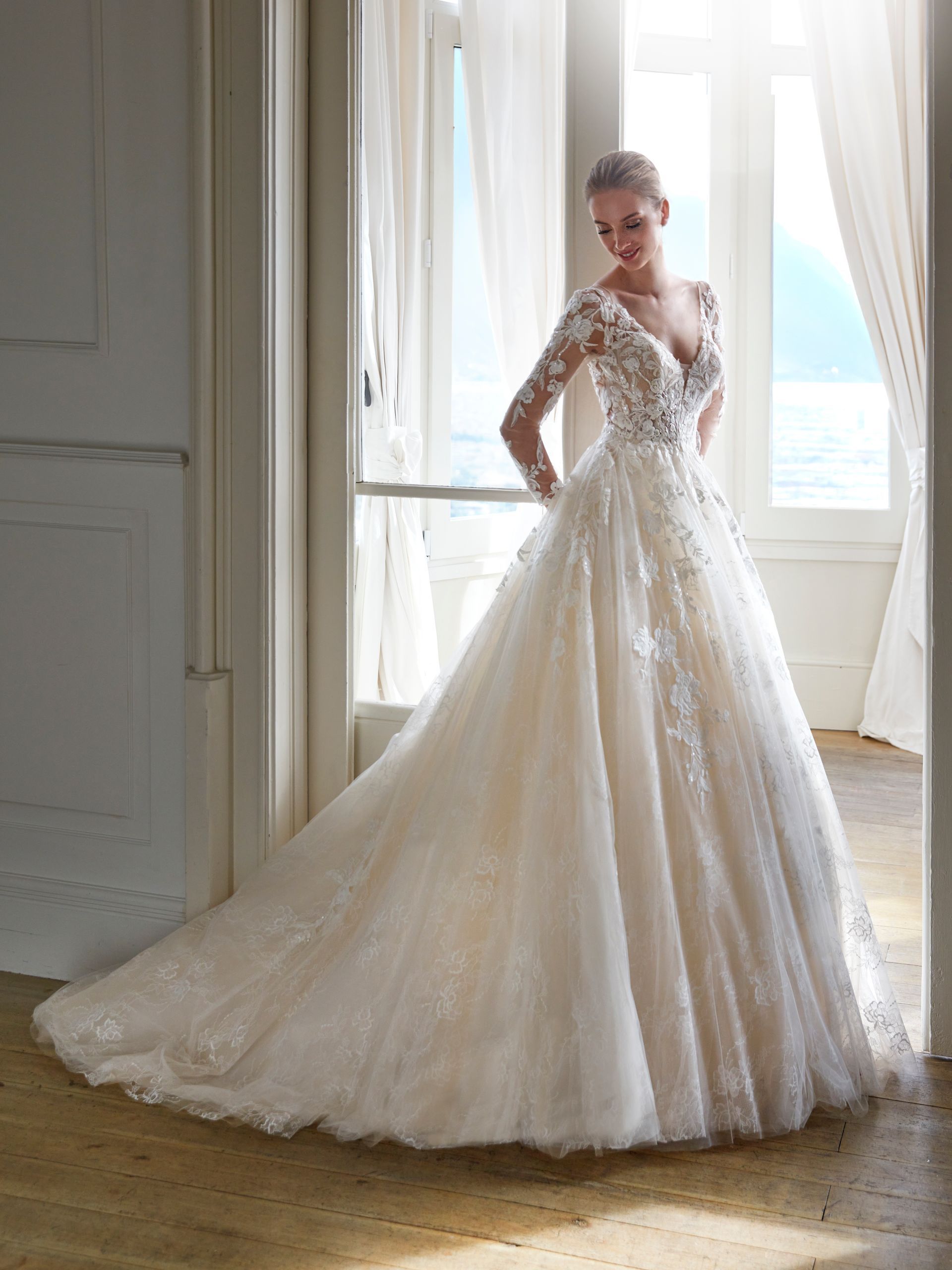 Donna in abito da sposa bianco con lungo strascico, in posa davanti a una finestra.