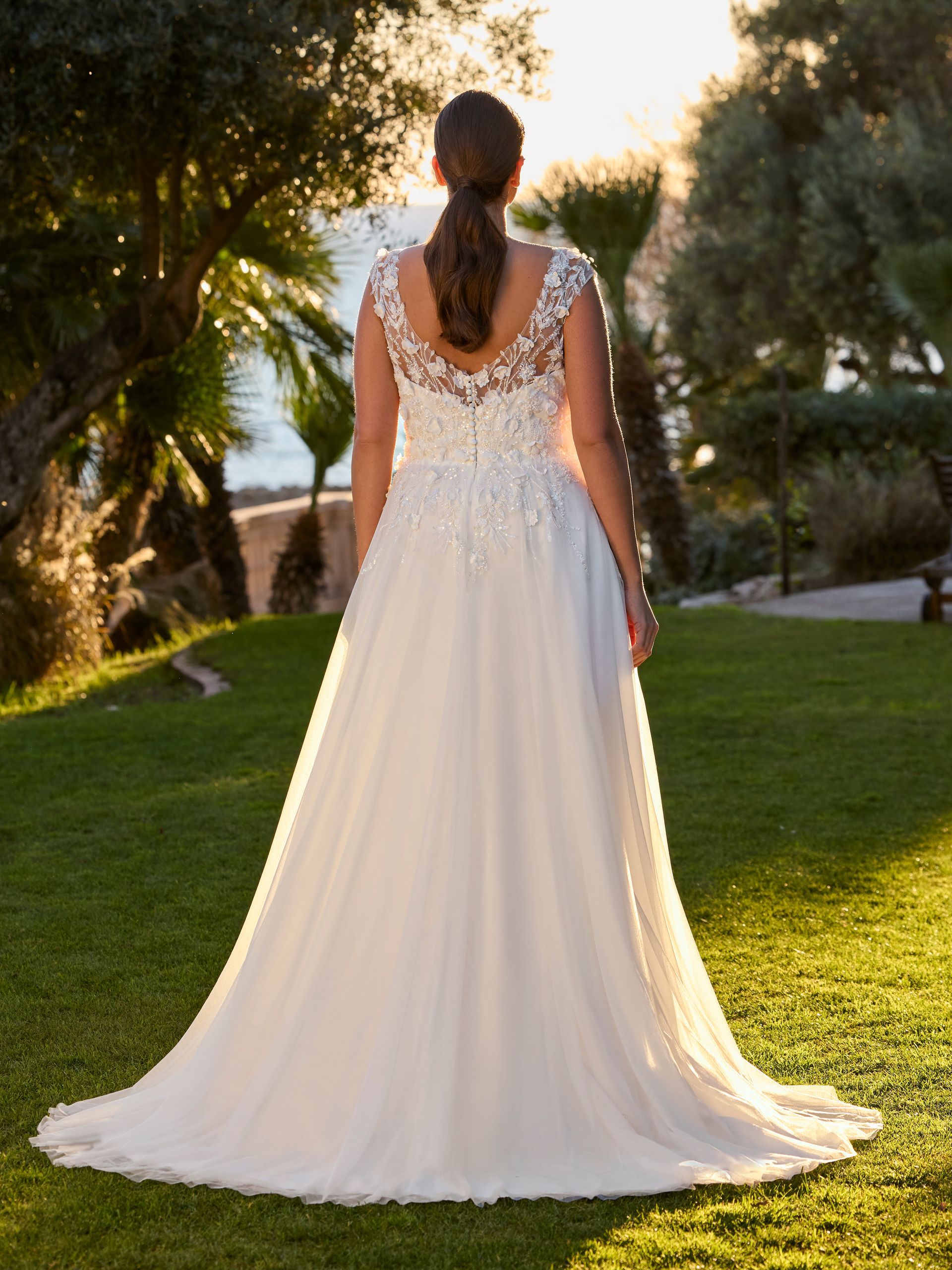 Donna in abito da sposa bianco, in piedi sull'erba, di spalle alla telecamera. La luce del sole splende, circondata da alberi verdi.