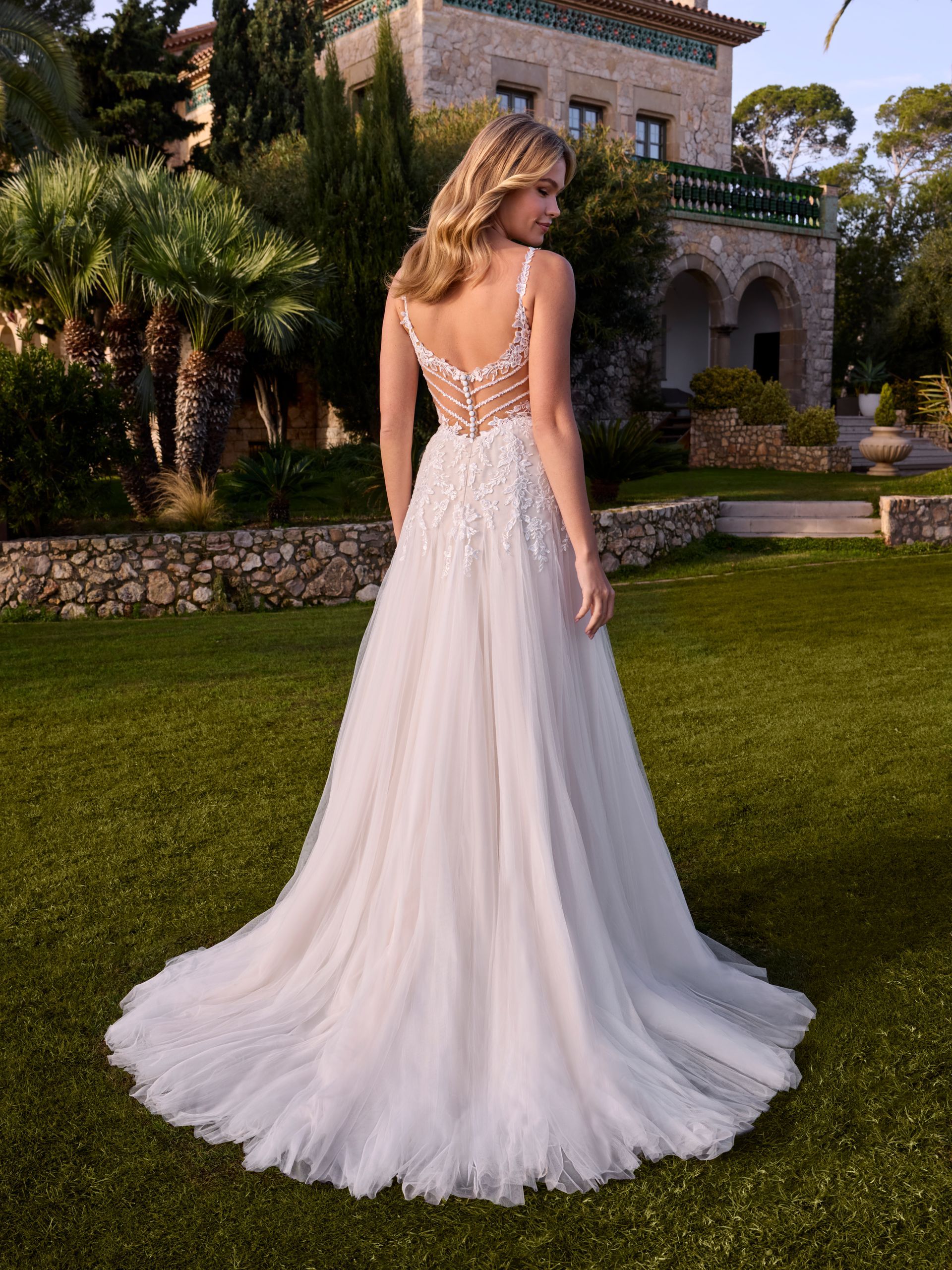 Donna in abito da sposa bianco con lungo strascico, in piedi in un giardino.