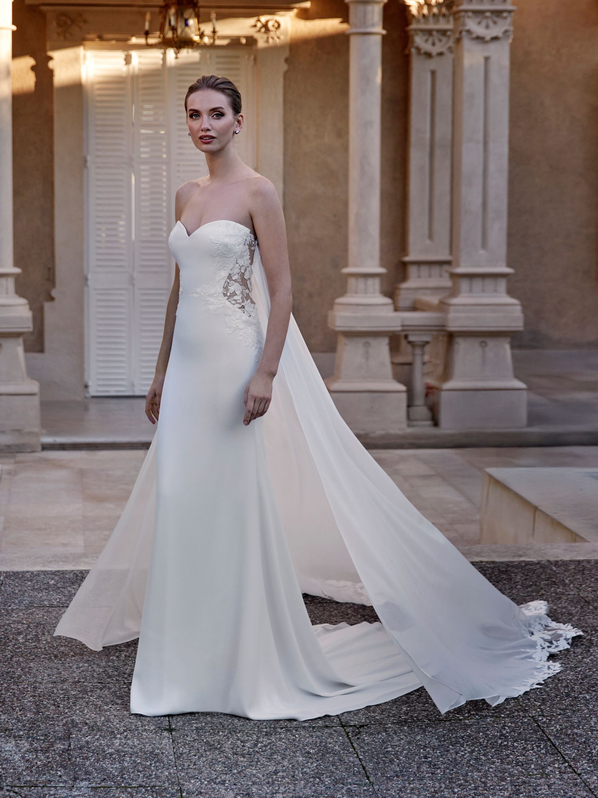 Donna in un abito da sposa bianco senza spalline con lungo strascico, in piedi all'esterno.