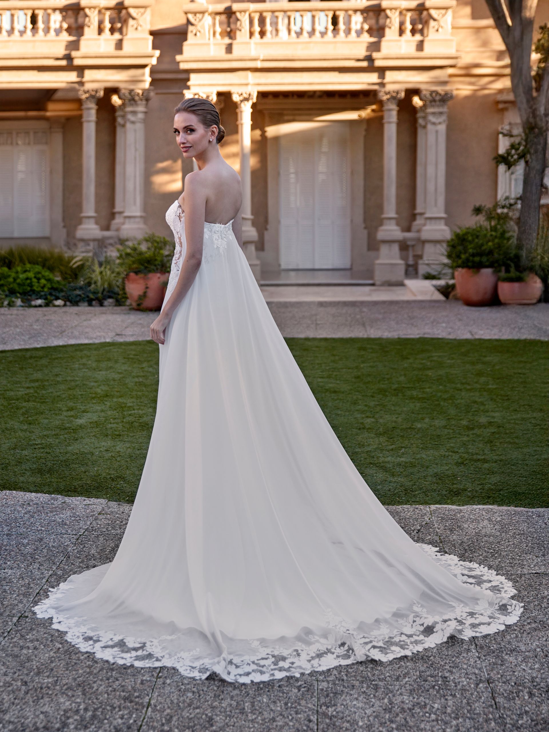 Donna in abito da sposa bianco con lungo strascico, in piedi su un sentiero di pietra all'aperto.