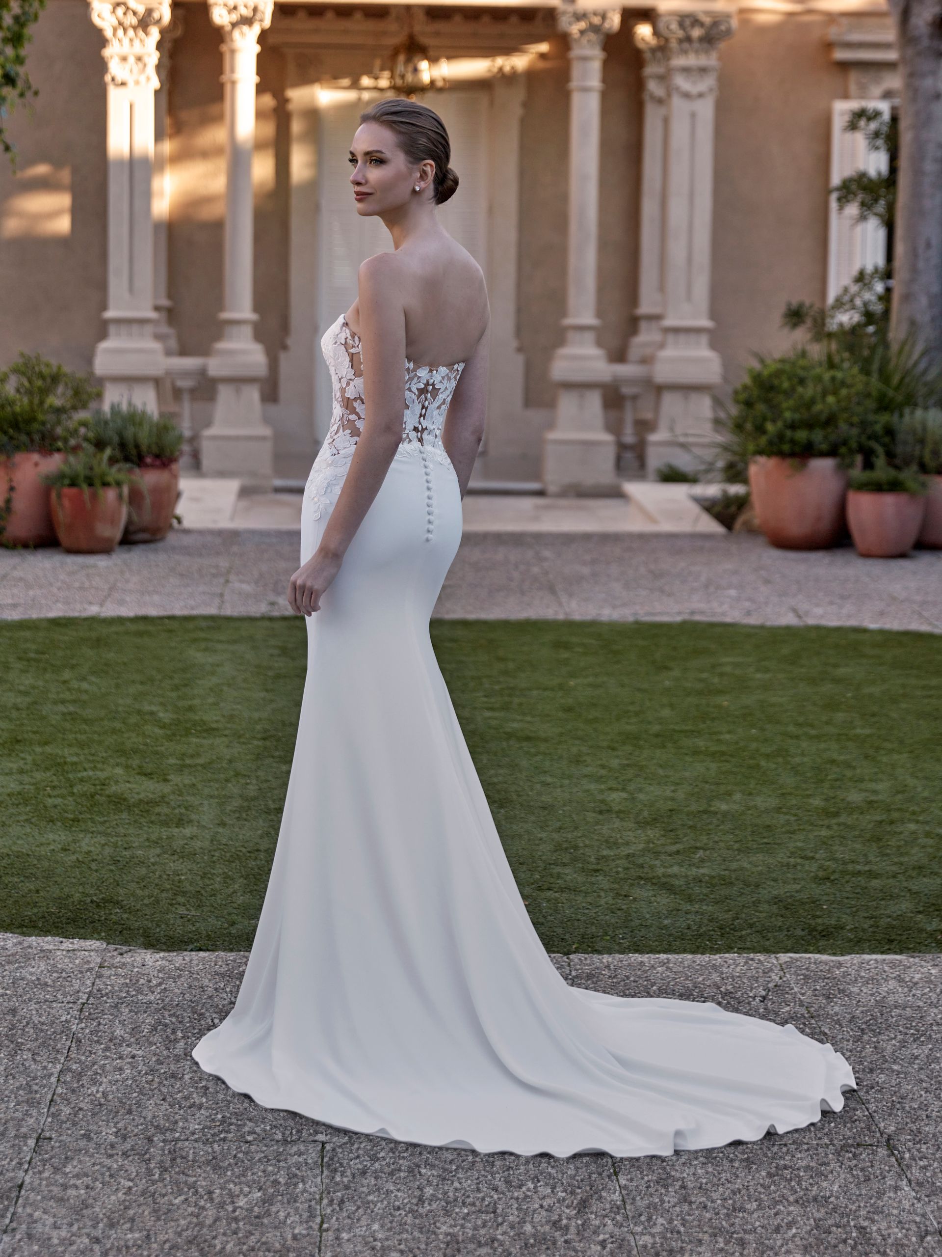 Una donna in abito da sposa bianco è in piedi sul sentiero, guardando oltre la sua spalla, nel giardino.