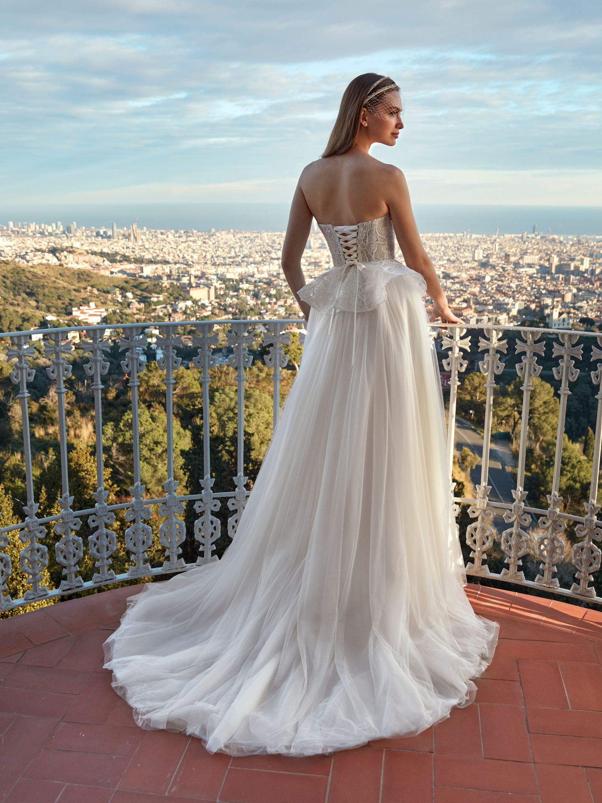 Sposa in abito bianco senza spalline con strascico, che guarda la città da un balcone.
