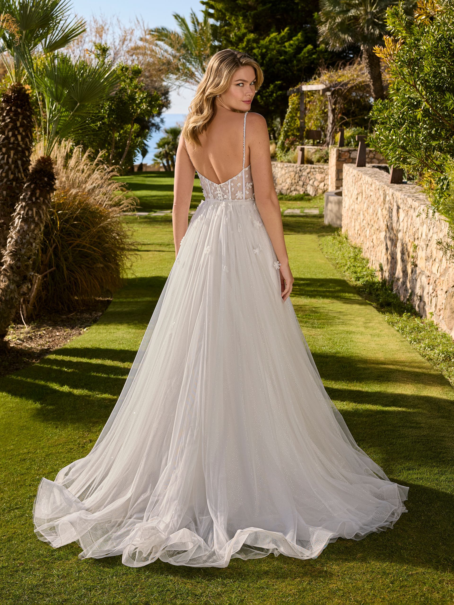 Una donna con un abito da sposa bianco con spalline sottili è in piedi in un giardino soleggiato.