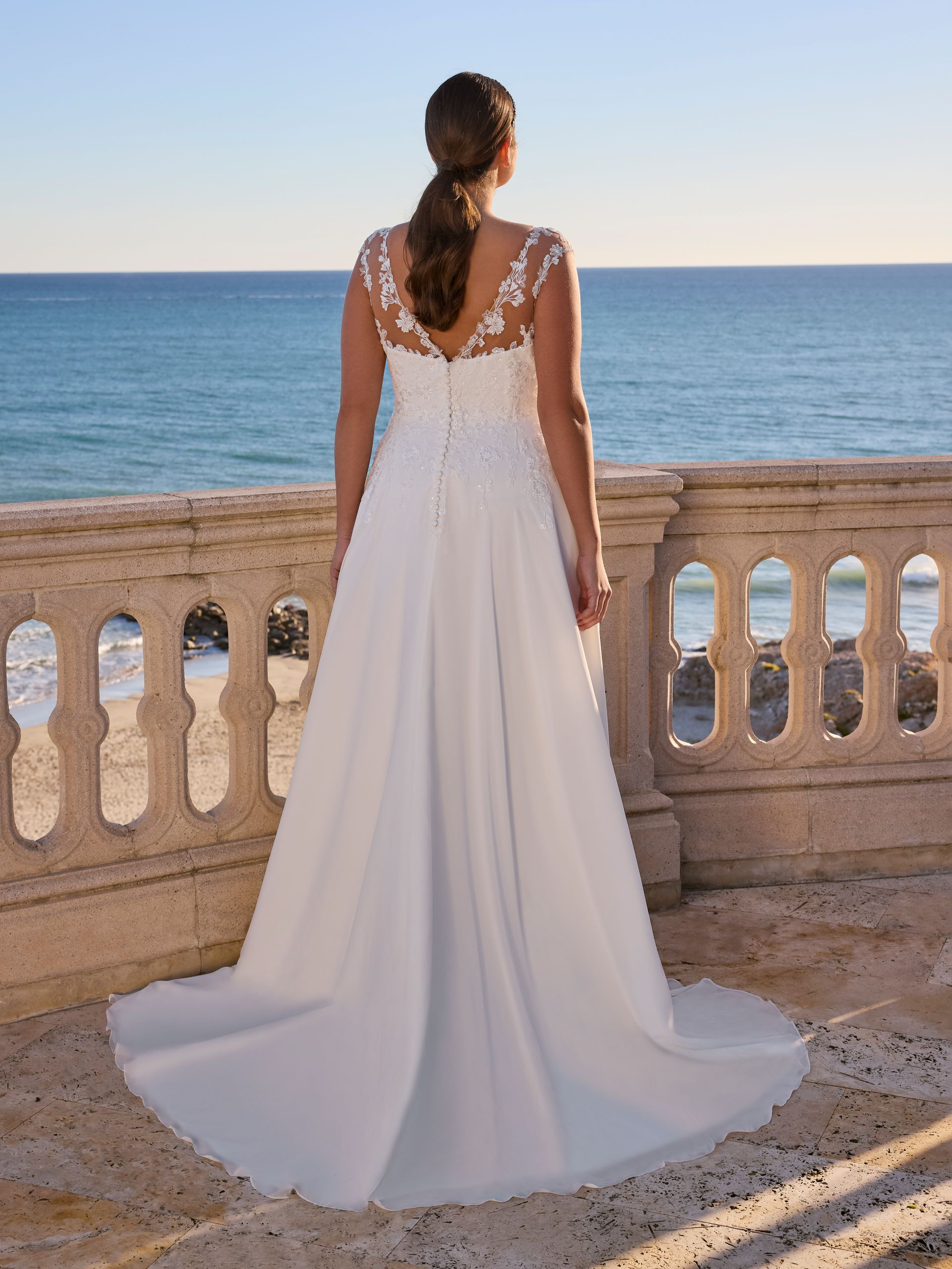 Una donna con un abito da sposa bianco, di spalle alla telecamera, è in piedi su un balcone con vista sull'oceano.