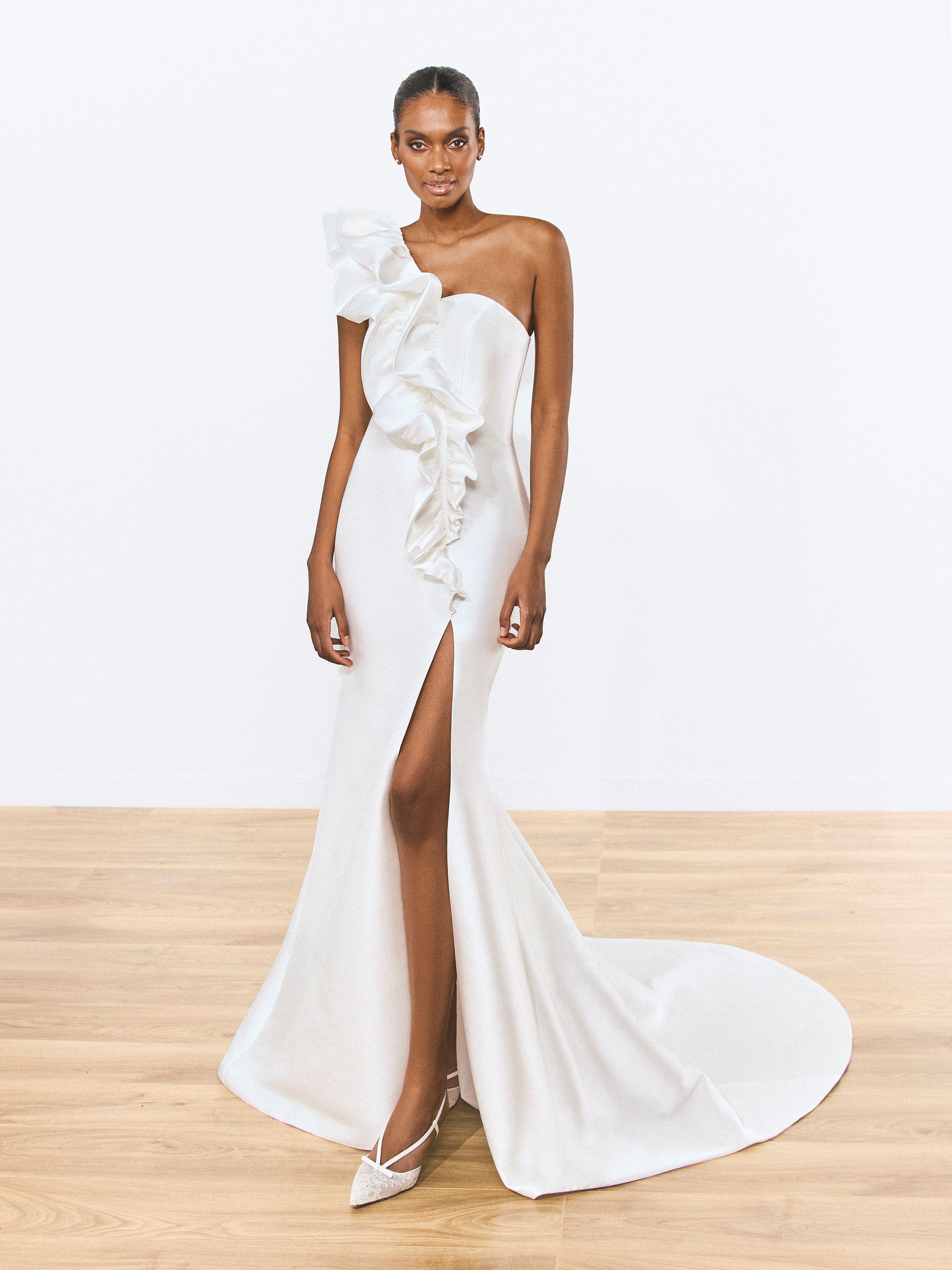 Donna in abito bianco monospalla con corpetto arricciato, spacco profondo e strascico, in piedi su un pavimento di legno.