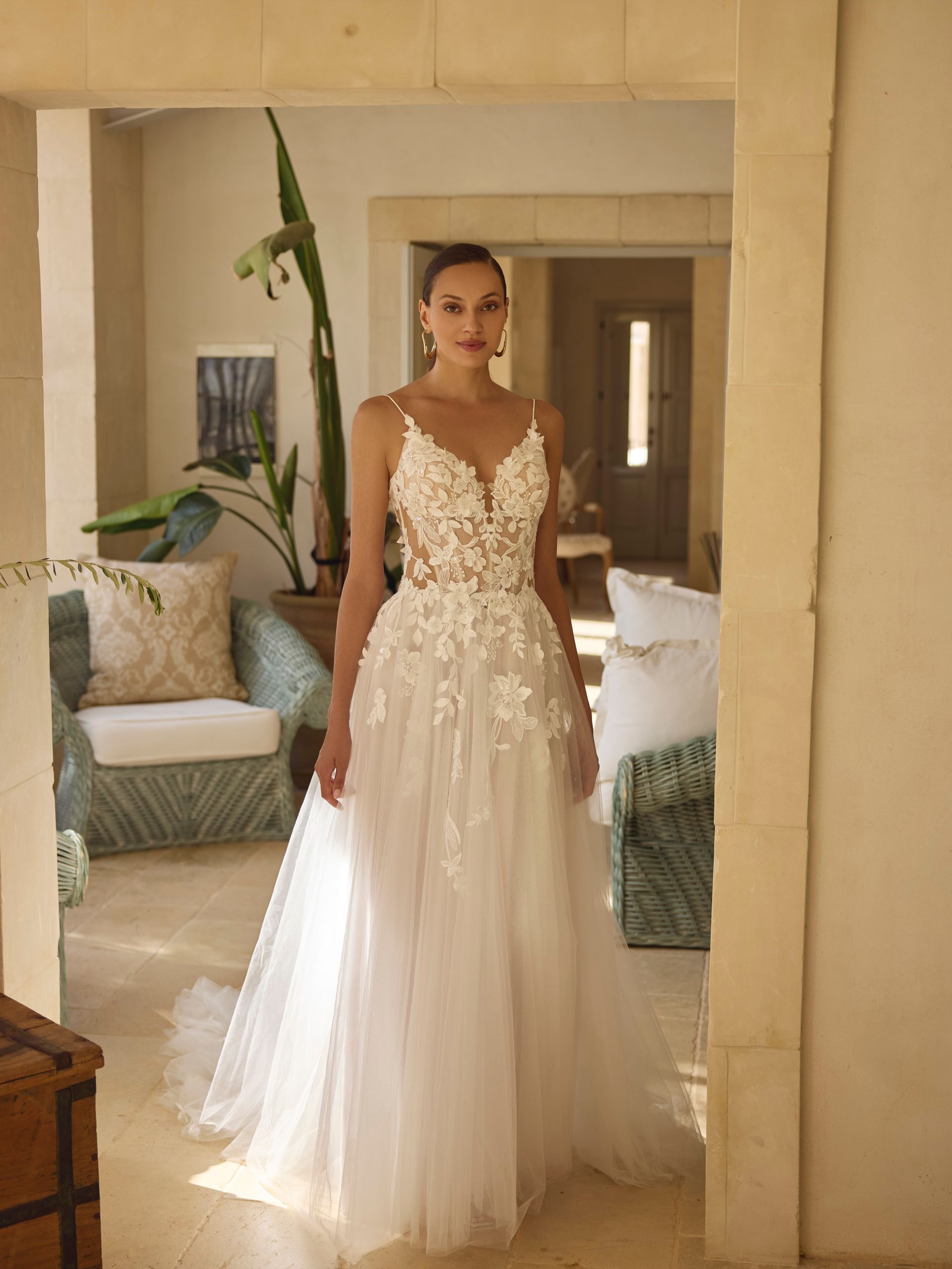 La sposa indossa un abito da sposa bianco a fiori ed è in piedi davanti a una porta aperta, in un interno luminoso, con luce naturale.