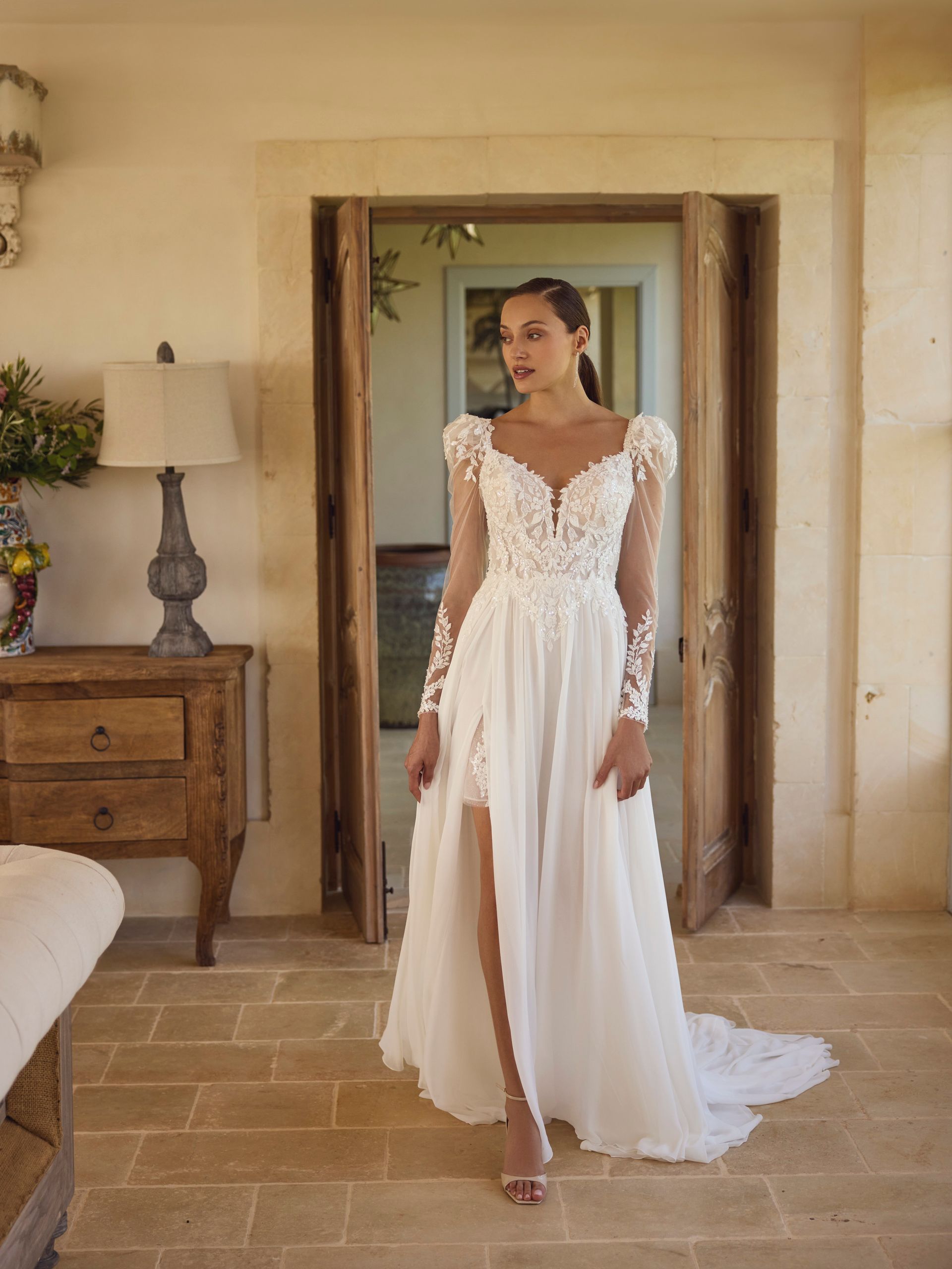 Donna in abito da sposa bianco con lunghe maniche in pizzo e spacco profondo, mentre attraversa la porta.