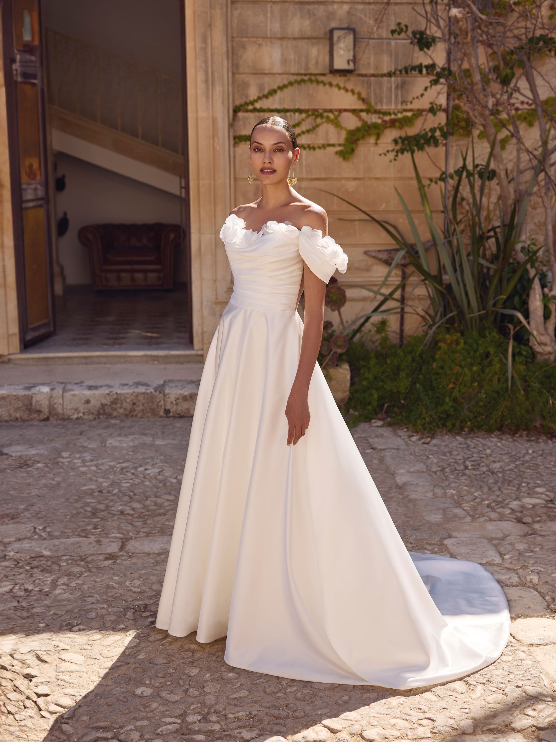 Una donna con un abito da sposa bianco a spalle scoperte posa davanti a un edificio in pietra.