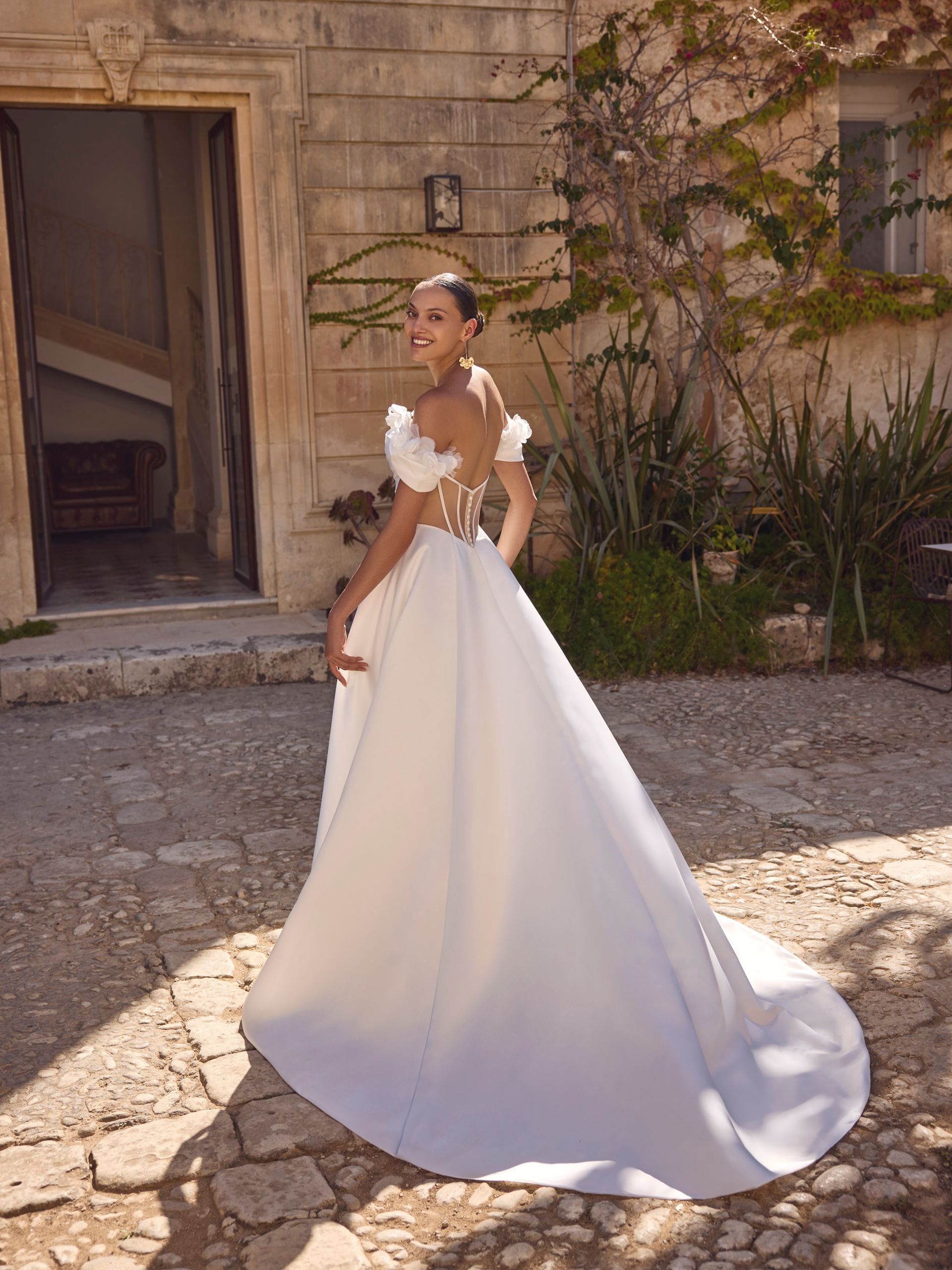 Una donna in abito da sposa bianco con lungo strascico posa all'aperto davanti a un edificio in pietra.