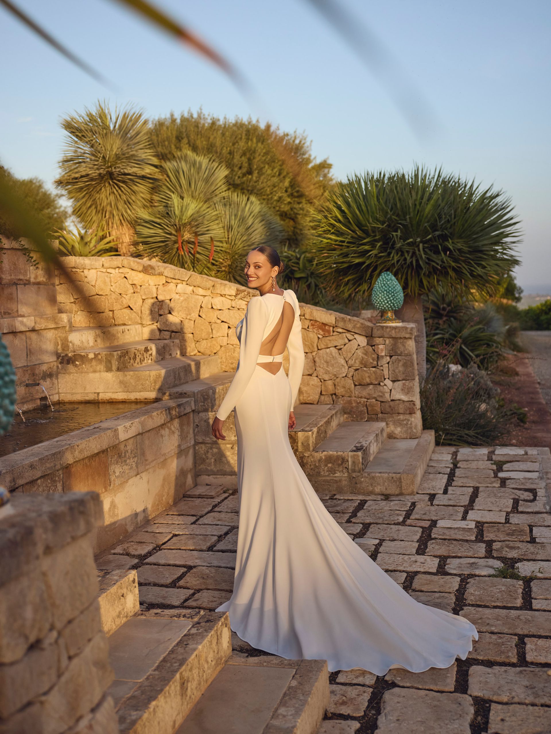 Donna in abito bianco su gradini di pietra, schiena scoperta, lungo strascico, ambientazione all'aperto.