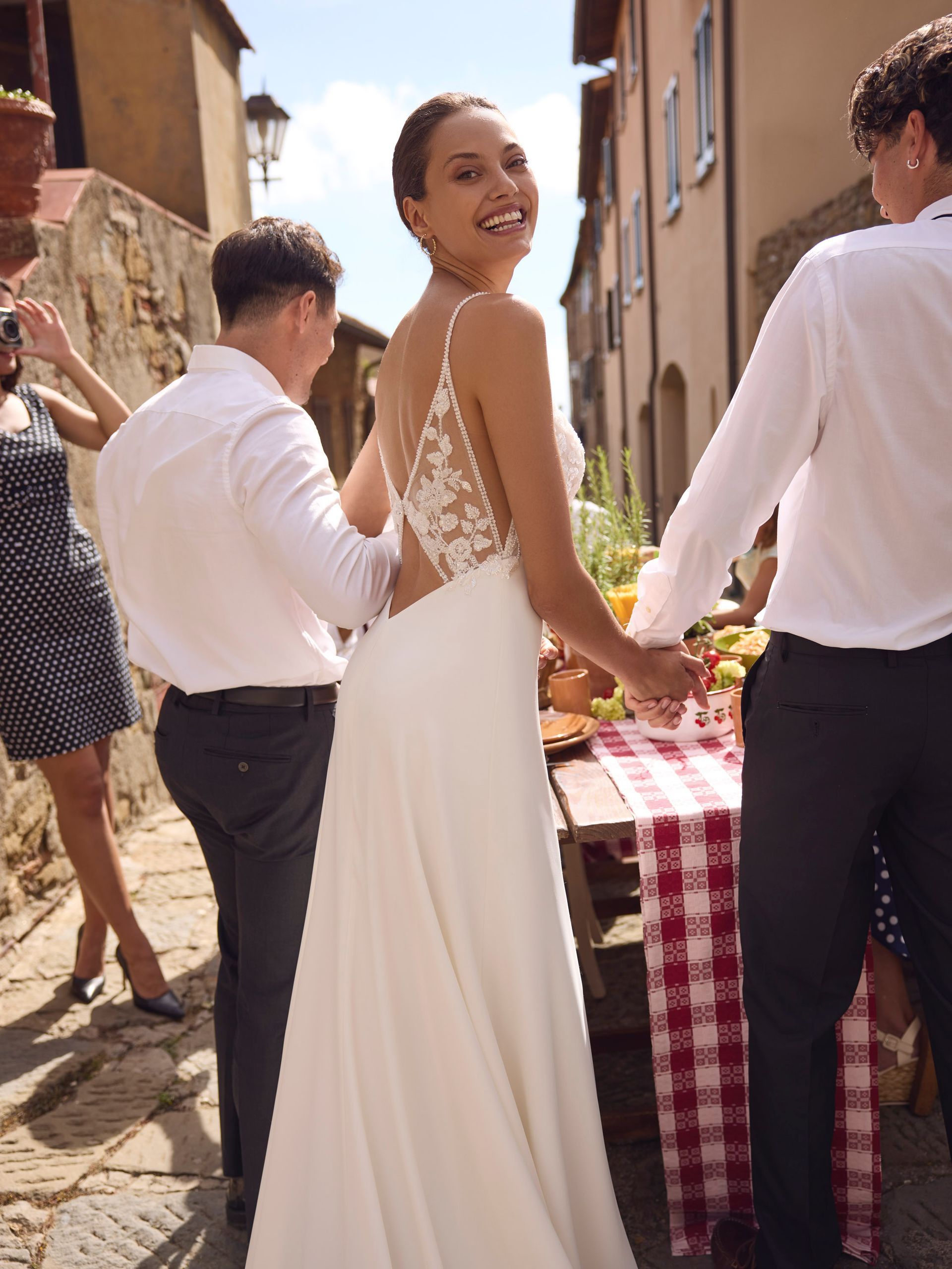 Una donna in abito bianco sorride, tenendo per mano due uomini vicino a un tavolo con del cibo; ambiente italiano all'aperto.