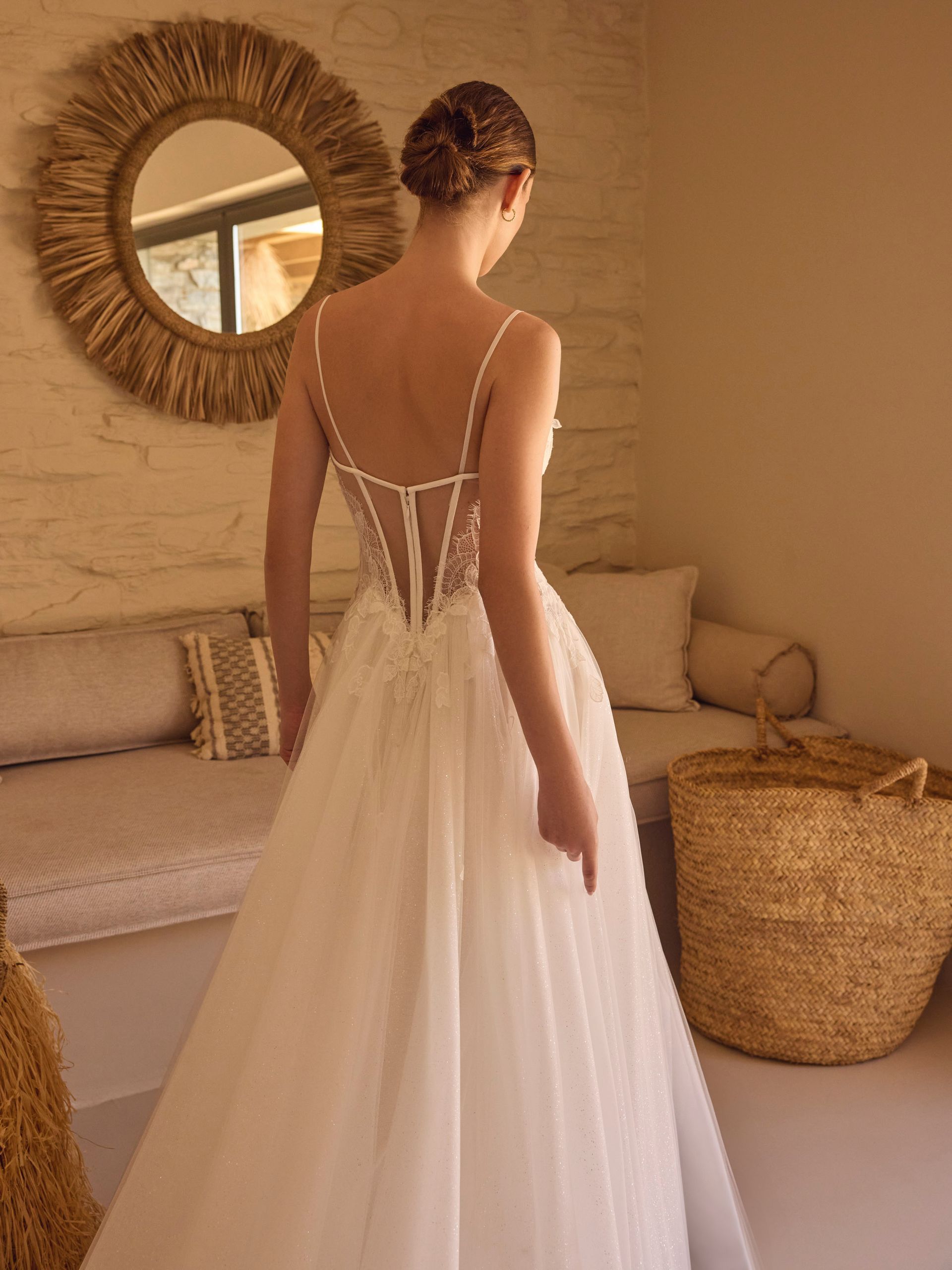 Donna in abito da sposa bianco, di spalle, in piedi in una stanza dai toni neutri.