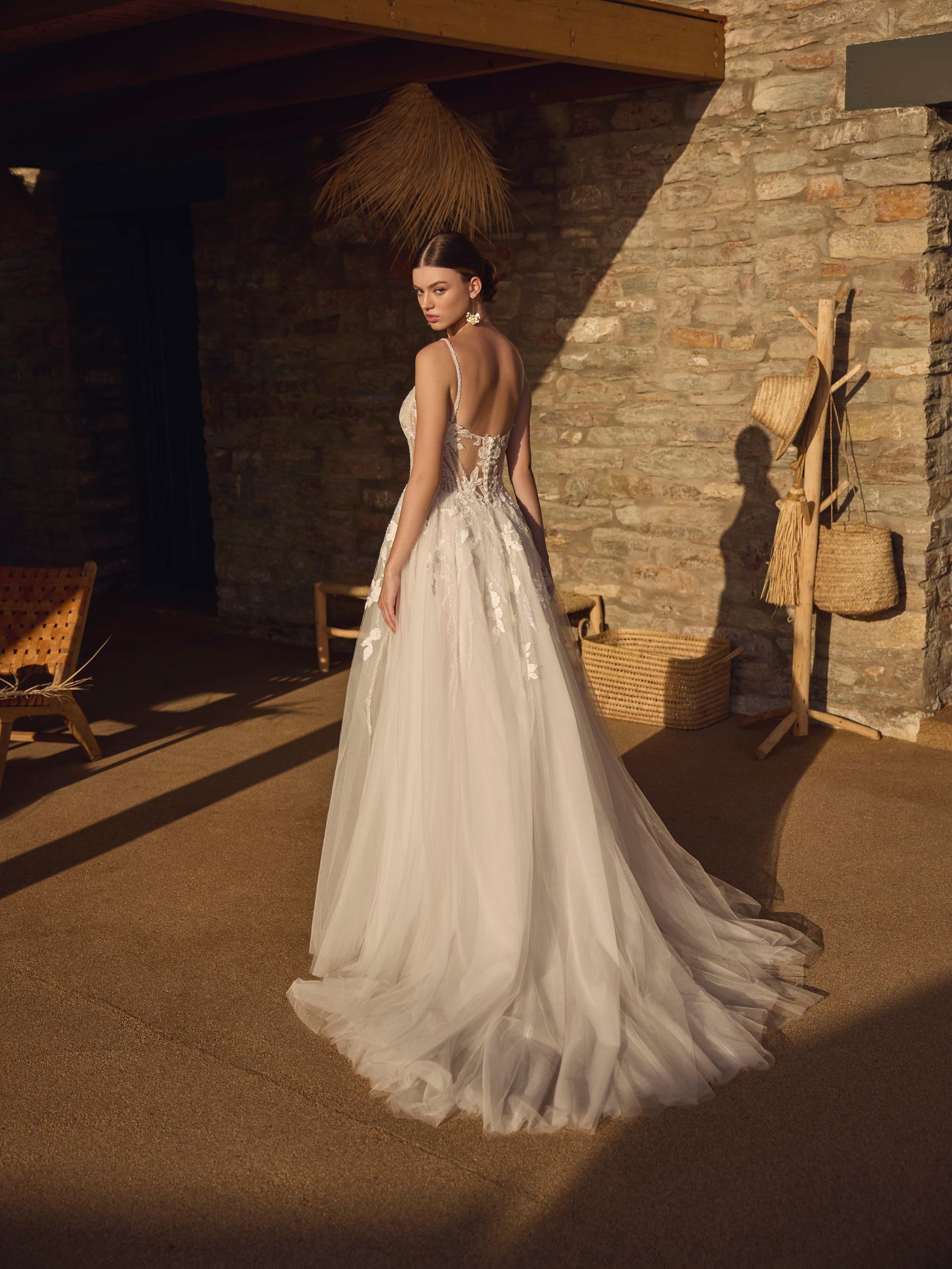 Donna in abito da sposa bianco con strascico, all'aperto vicino a un muro di pietra, che guarda oltre la spalla.