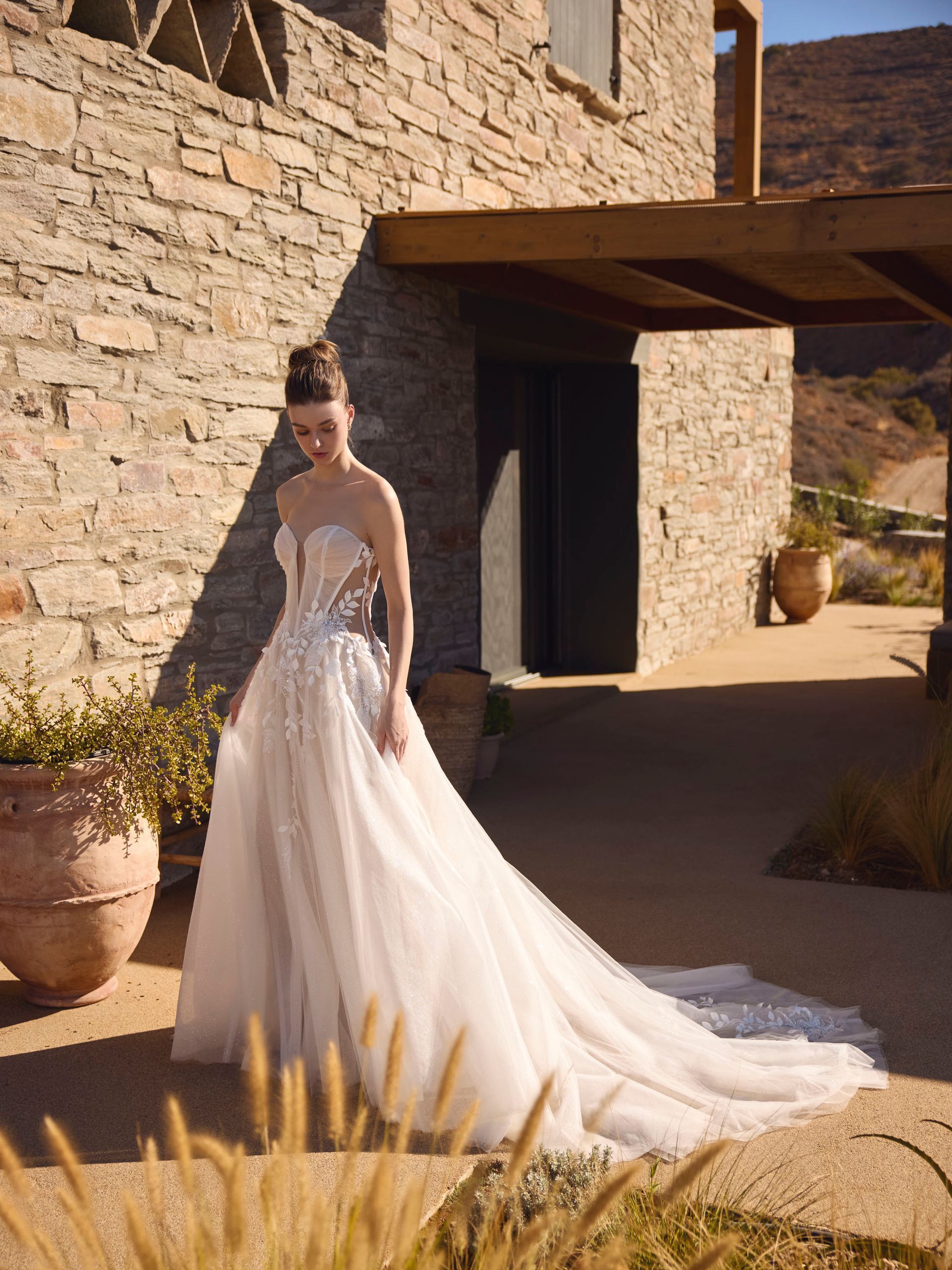 Una donna in abito da sposa bianco posa davanti a un edificio in pietra, con uno strascico che scorre dietro.