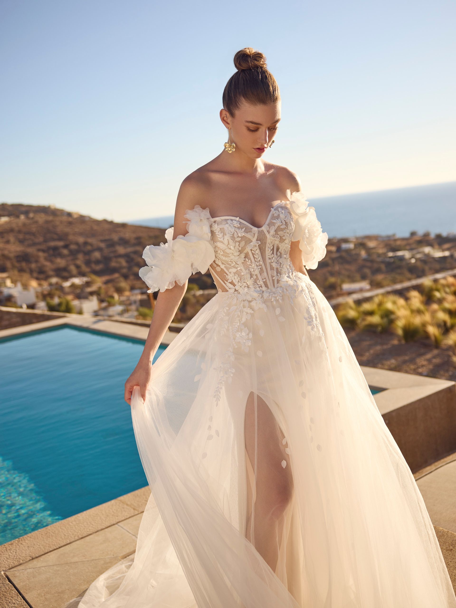 Donna in abito da sposa bianco, all'aperto vicino alla piscina, spacco profondo, spalle scoperte, acconciatura raccolta, orecchini.