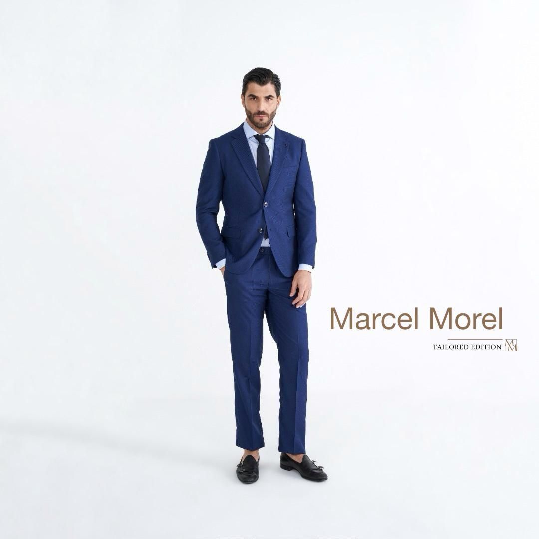 Uomo in abito blu con mano in tasca, sfondo bianco, logo Marcel Morel.