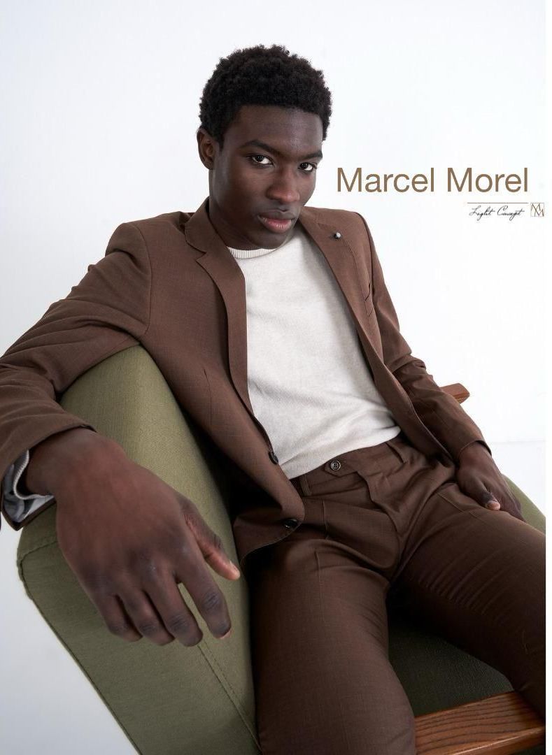 Uomo in abito marrone seduto su una sedia verde, che guarda lo spettatore.