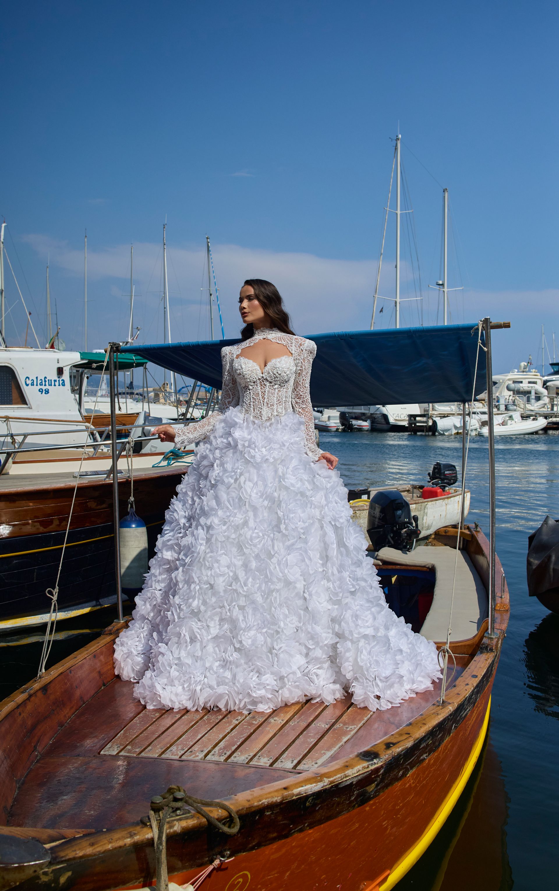 Una donna in abito da sposa bianco è in piedi su una piccola barca in un porto turistico, in una giornata di sole.