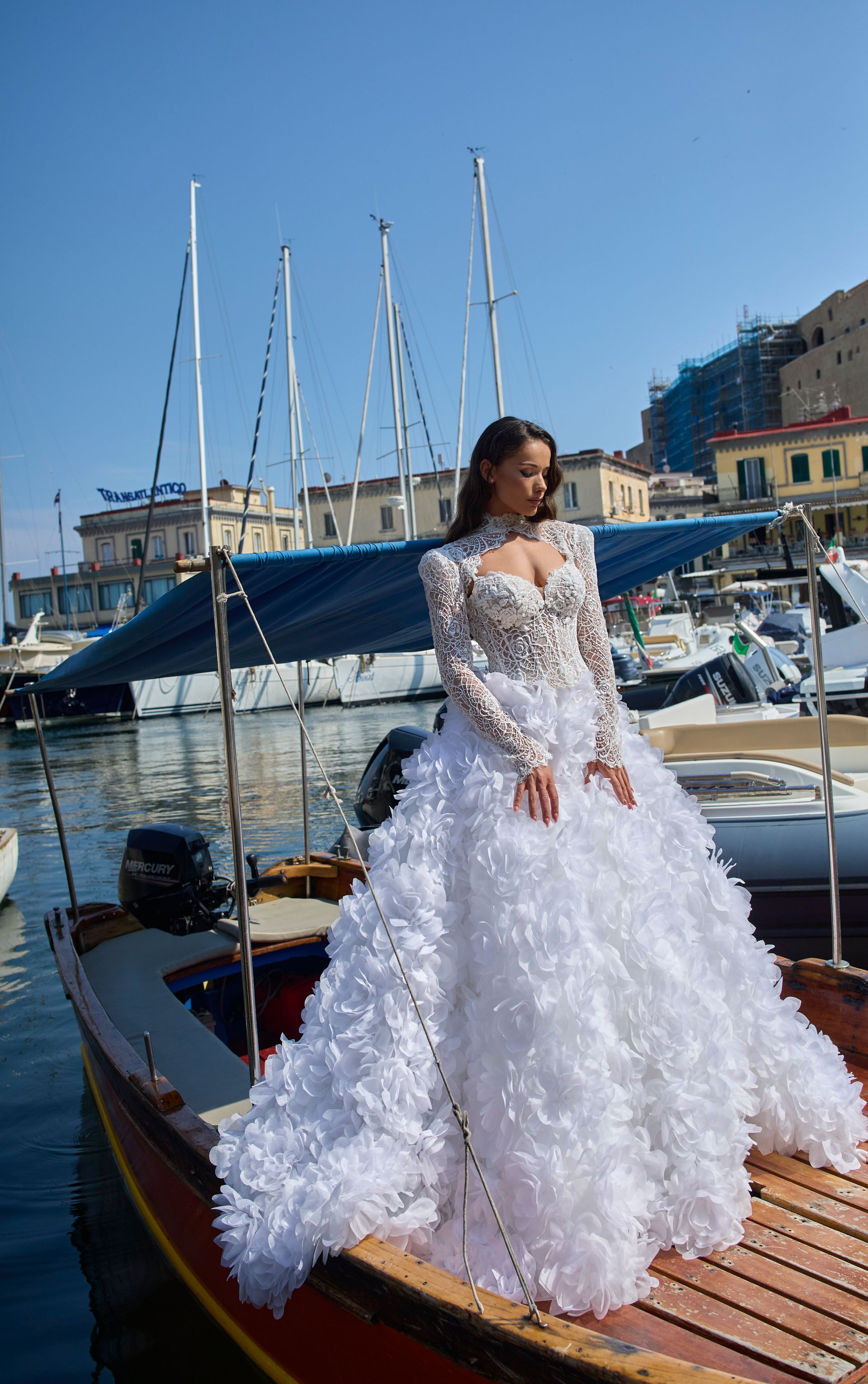Donna in abito bianco con volant su una barca in un porto, acqua blu, barche a vela ed edifici sullo sfondo.