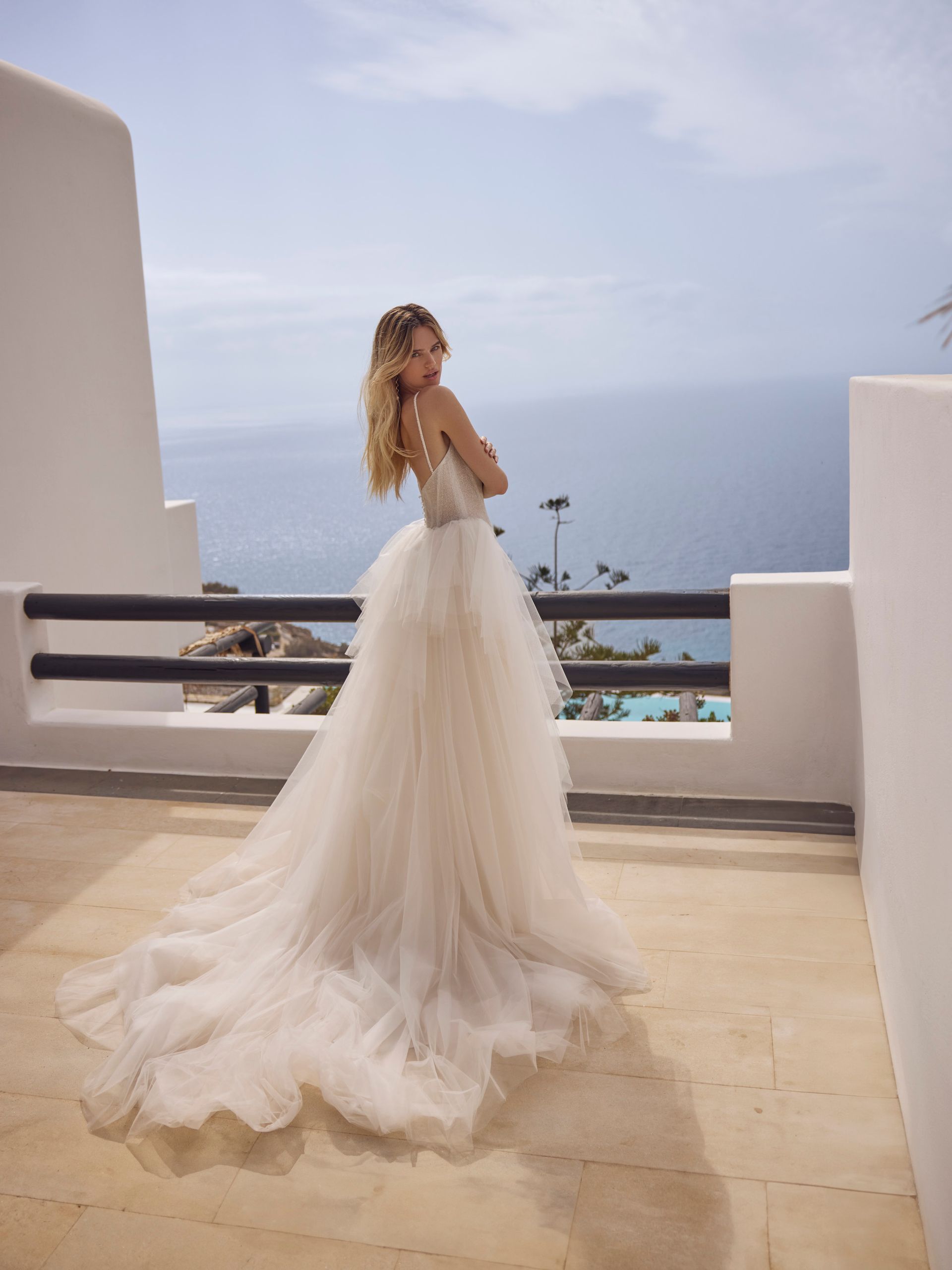 Donna bionda in abito da sposa bianco con lungo strascico, in posa su un balcone con vista sull'oceano.