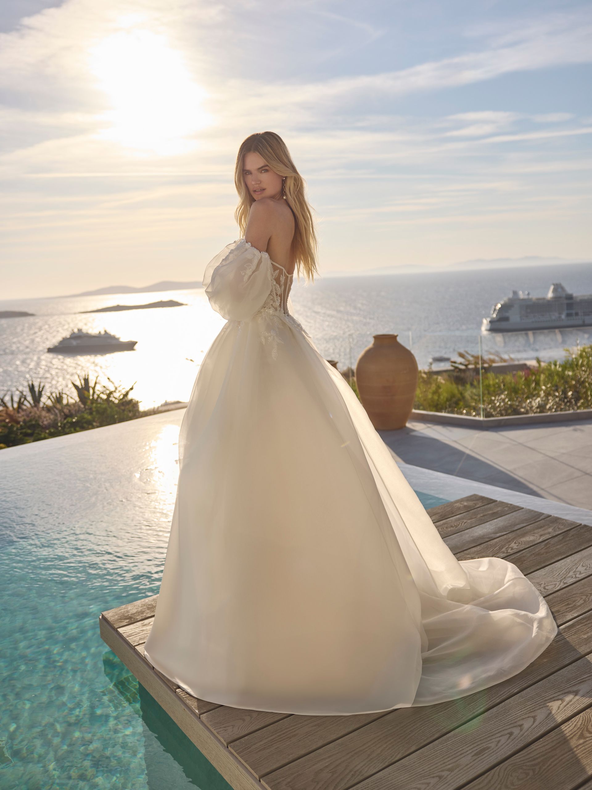 Una donna bionda in abito da sposa posa a bordo piscina con vista sull'oceano al tramonto.