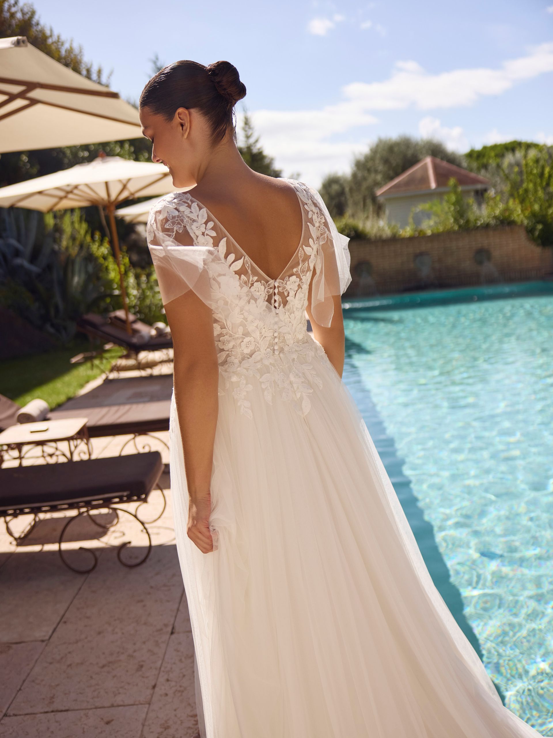 Donna in abito da sposa bianco vicino a una piscina, sorridente, all'aperto.