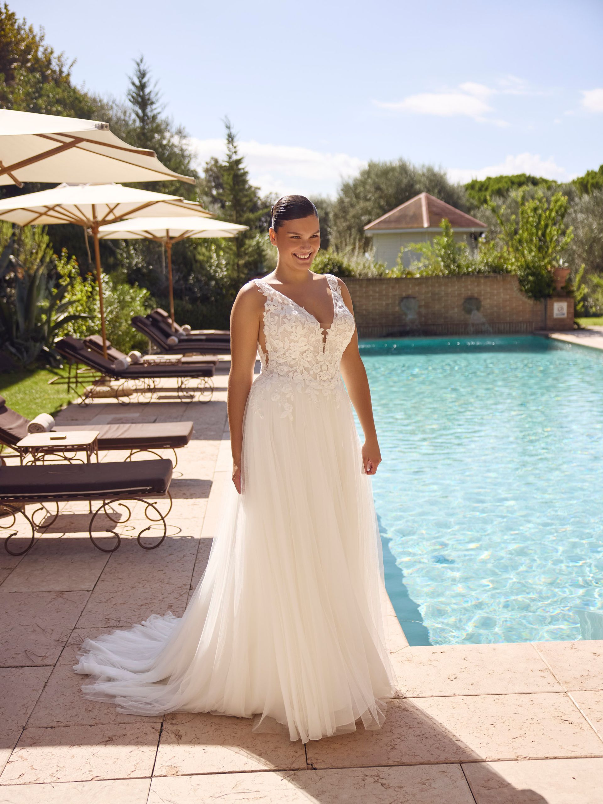 Una donna in abito da sposa bianco sorride a bordo piscina, in un ambiente soleggiato all'aperto.