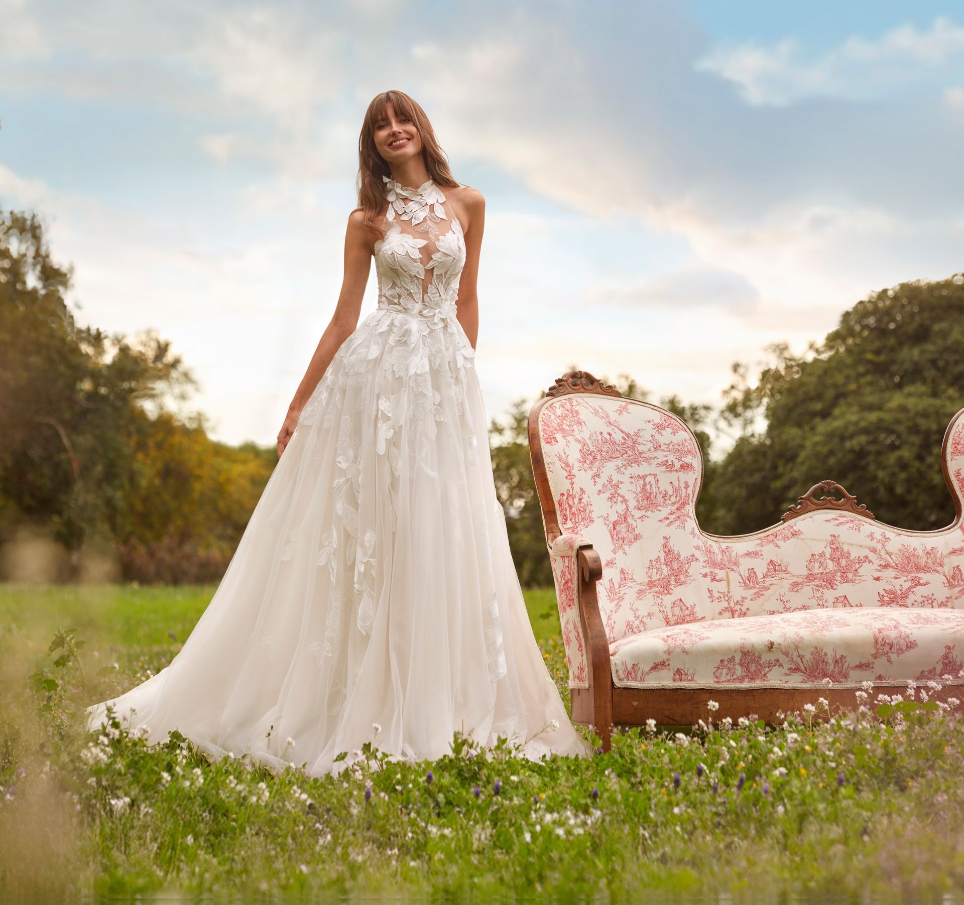 Una donna in abito da sposa bianco sorride in un campo, vicino a un divano floreale.