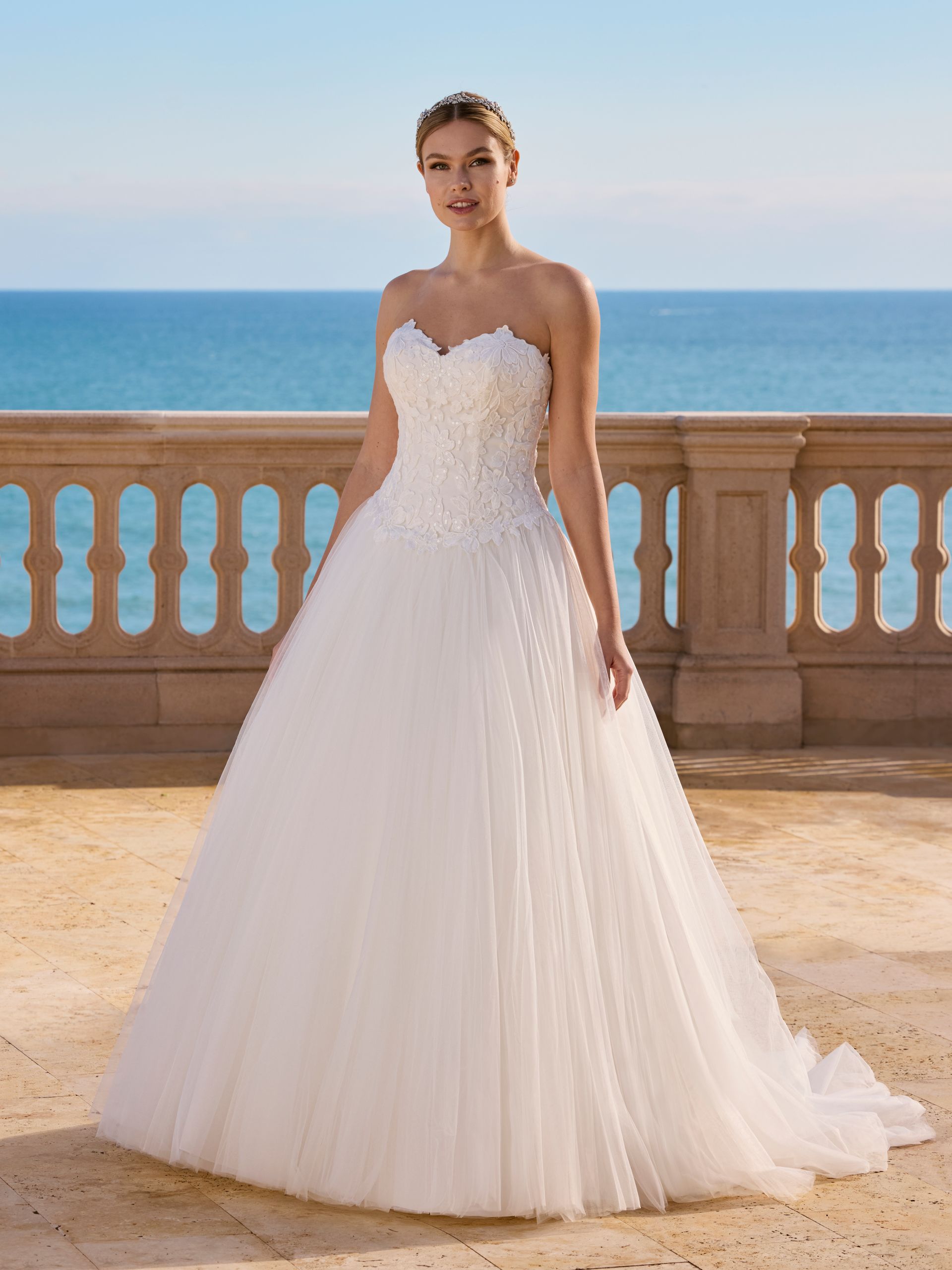Donna in abito da sposa bianco senza spalline, in posa su una terrazza con vista sull'oceano.