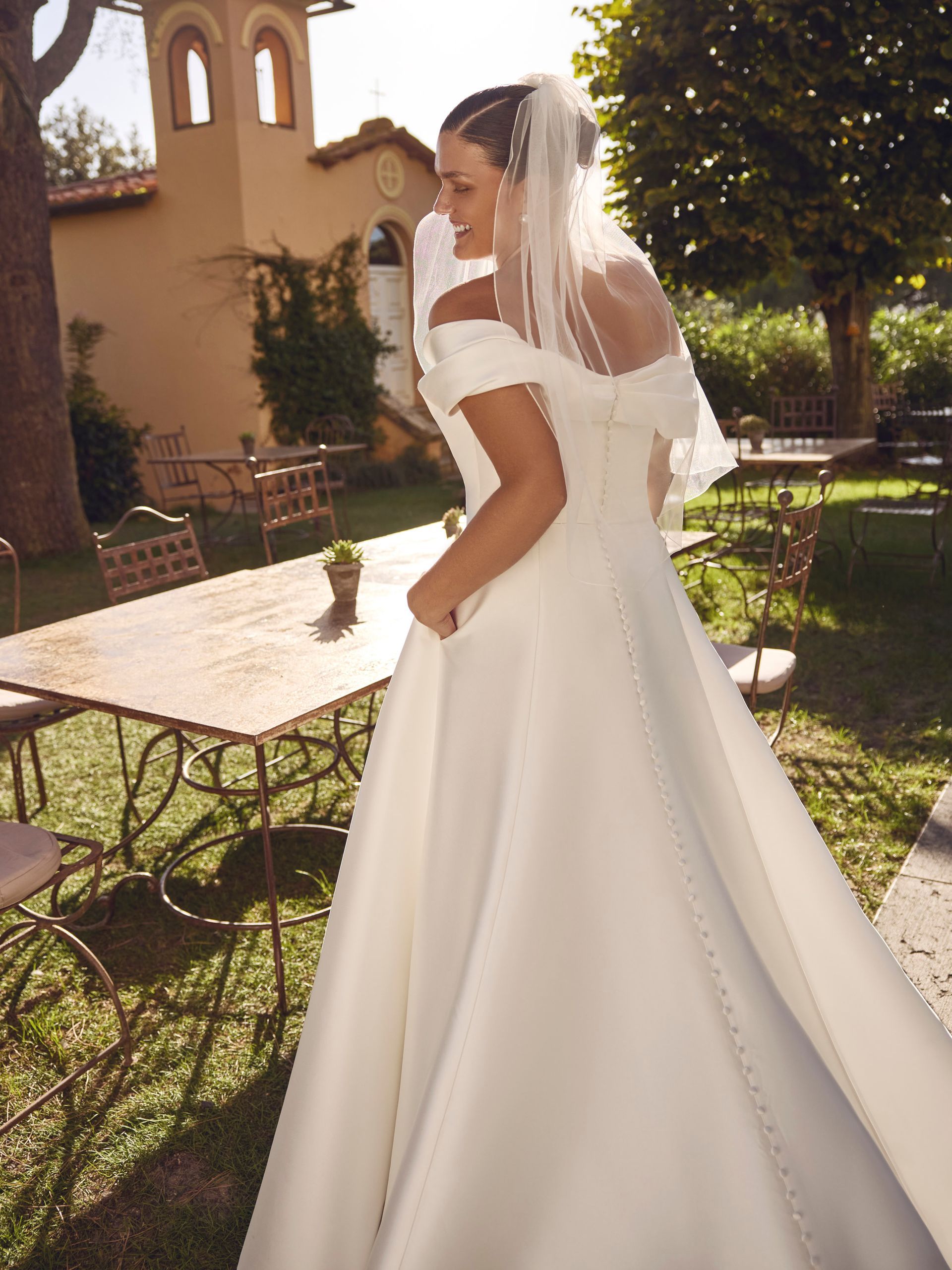 Sposa in abito bianco con spalle scoperte, velo, all'aperto, vicino al tavolo e in giardino.