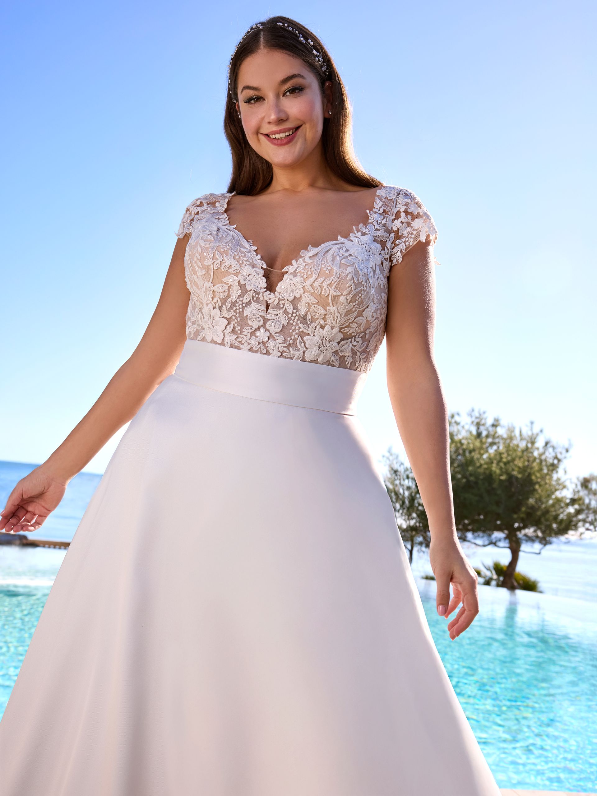 Donna in abito da sposa sorride, posa all'aperto vicino alla piscina; corpetto avorio, gonna bianca.