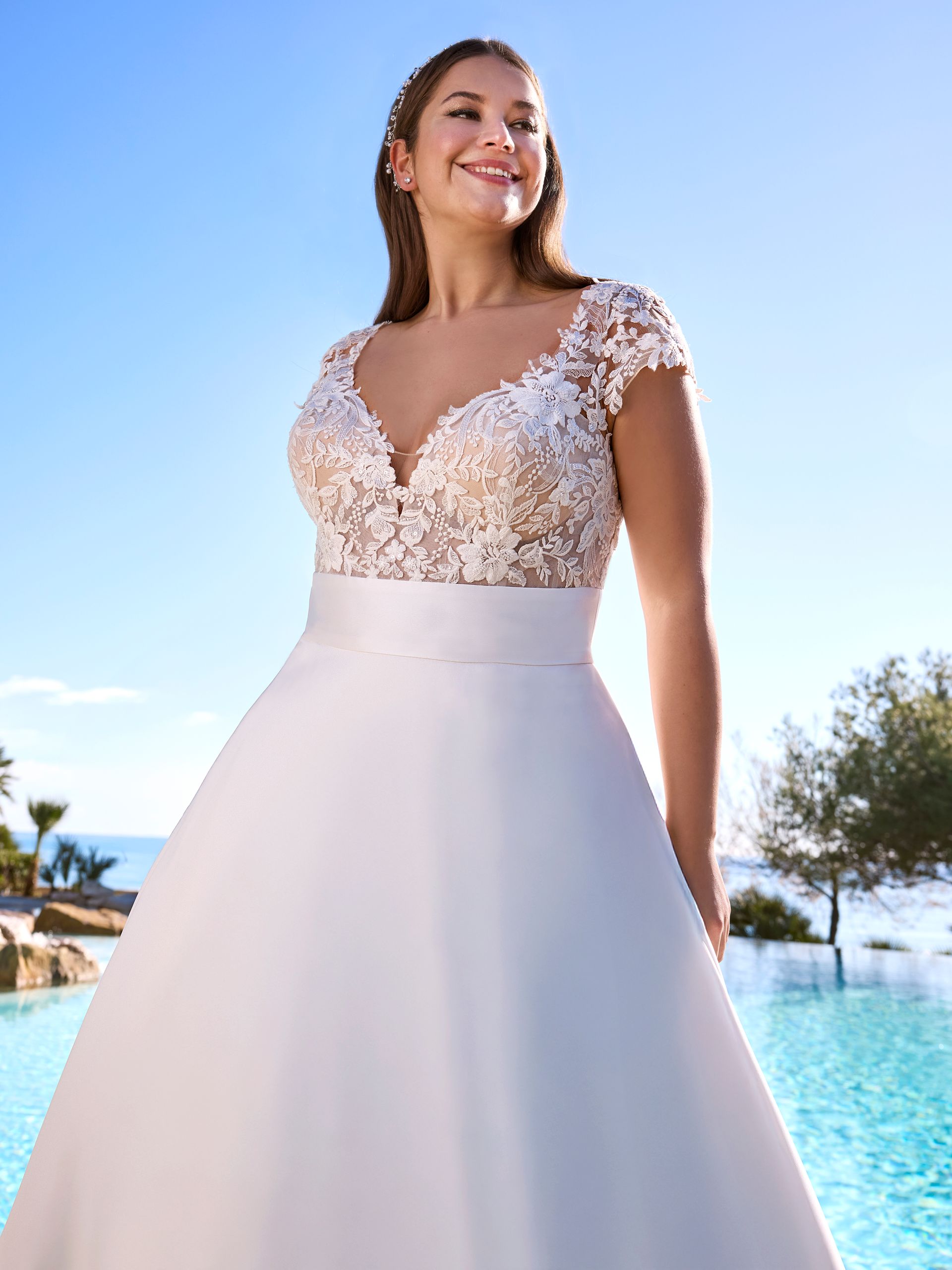 Donna in abito da sposa bianco, sorridente, all'aperto, a bordo piscina. L'abito ha un corpetto in pizzo e una gonna ampia.