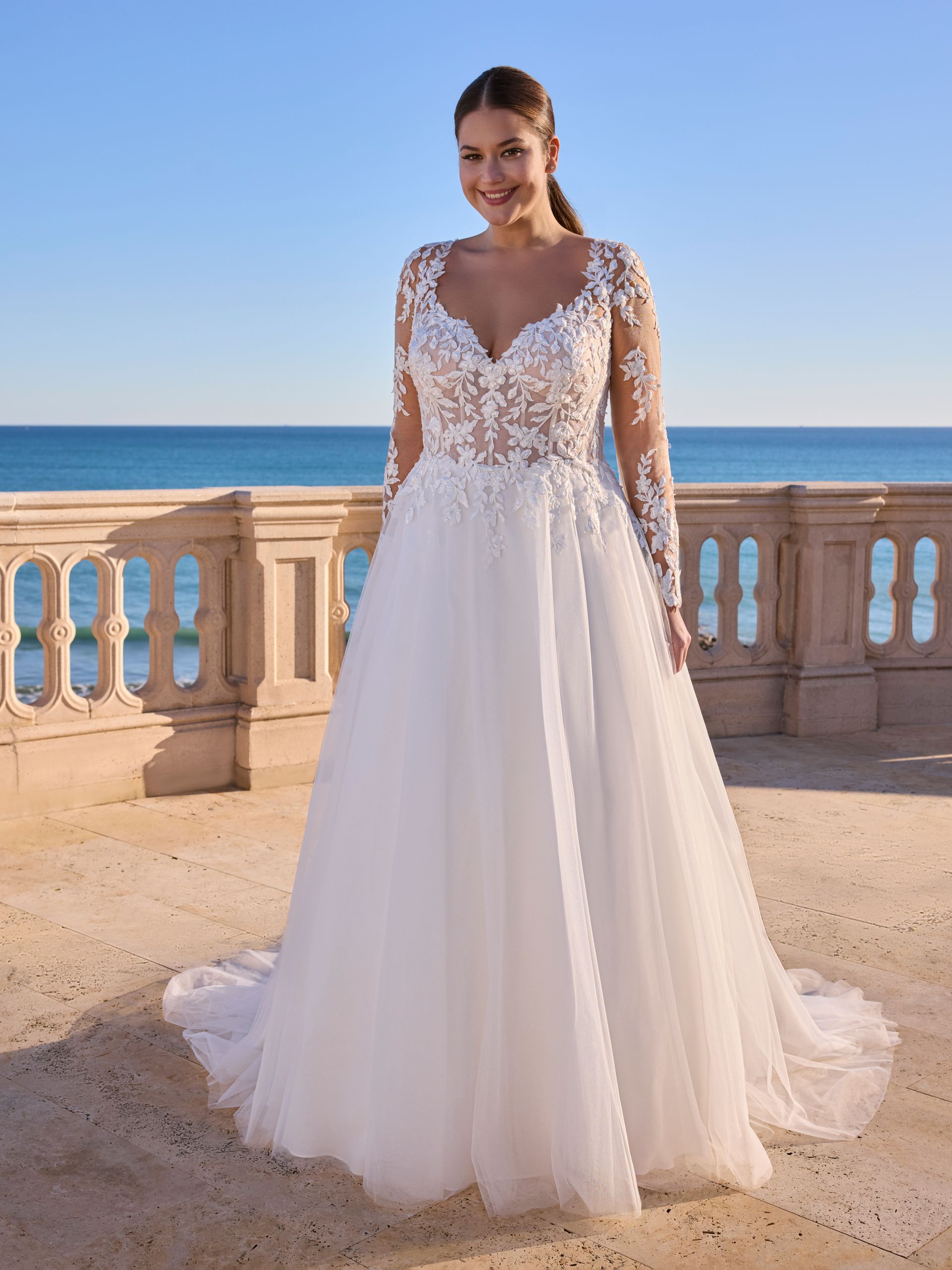 Una sposa con un abito bianco a maniche lunghe e corpetto floreale sorride su una terrazza con vista sull'oceano.