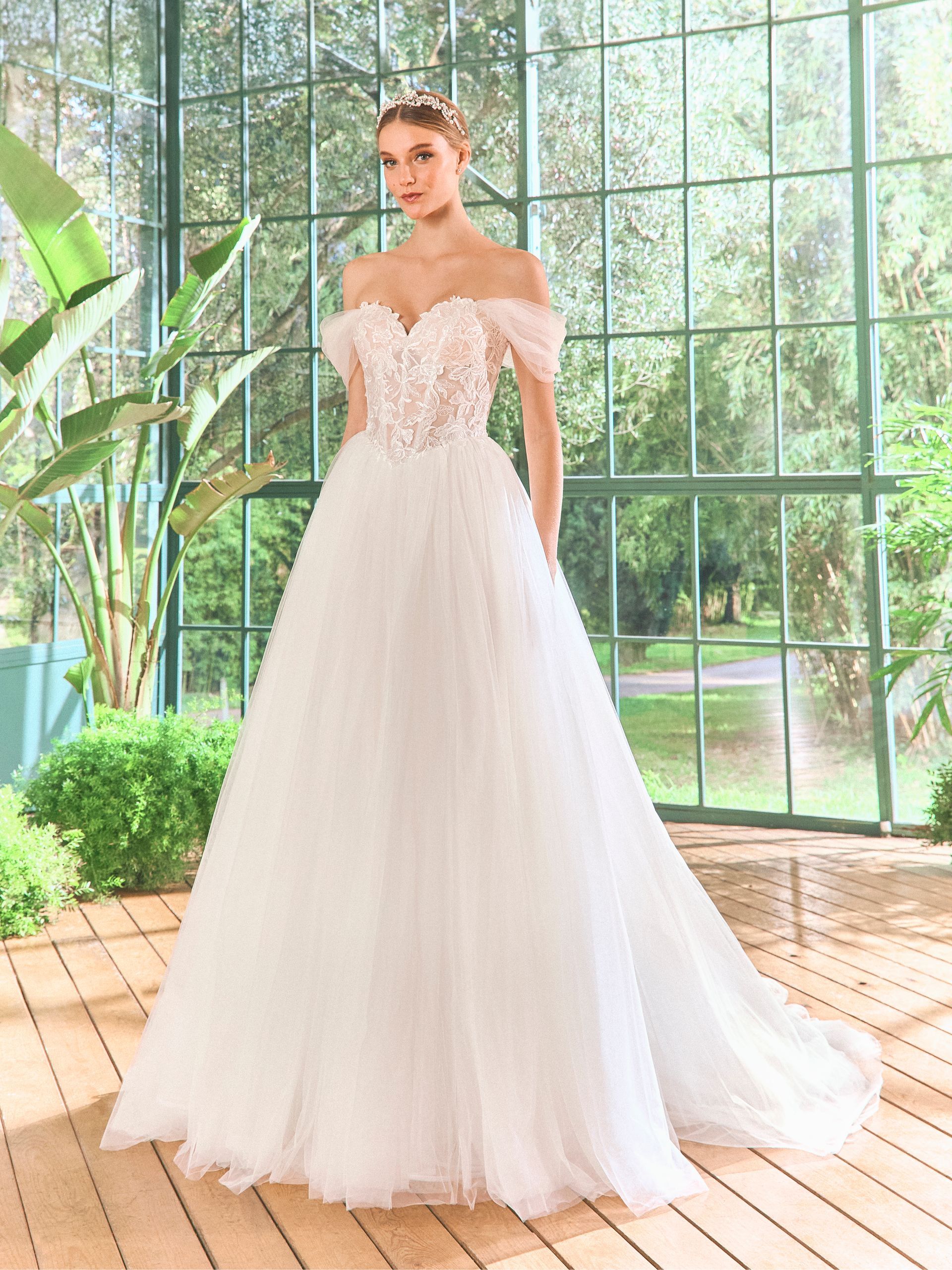 Donna in abito da sposa bianco con spalle scoperte e corpetto floreale, in piedi in una serra soleggiata.