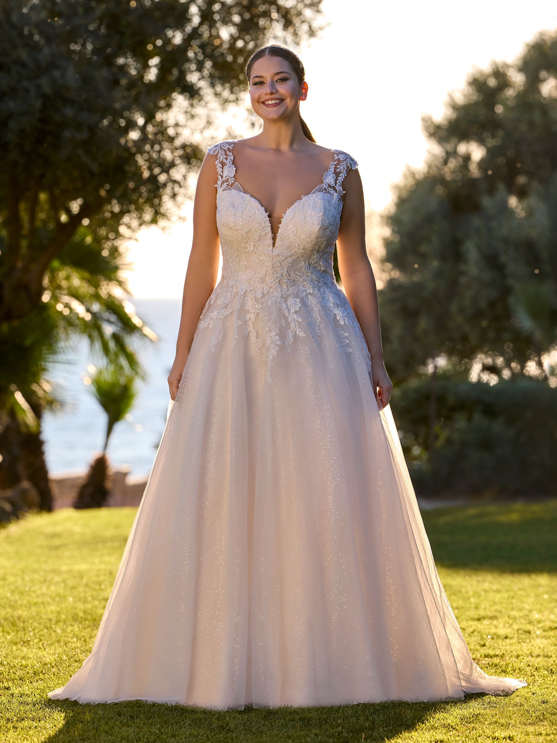 Donna in abito da sposa, sorridente, in piedi in un giardino soleggiato con una spiaggia sullo sfondo.