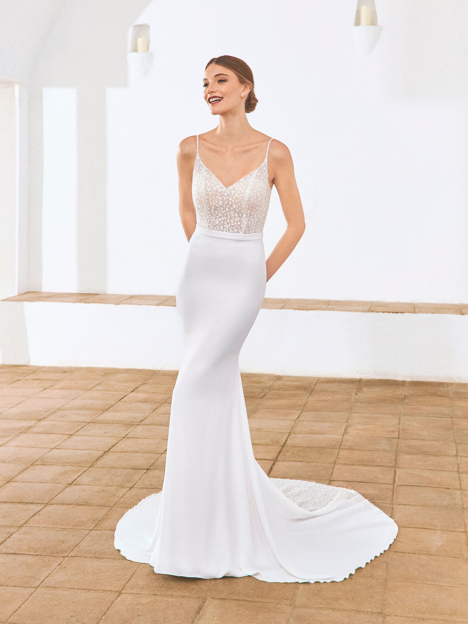 Una donna in abito da sposa bianco con corpetto di perline e gonna a sirena sorride, in casa.