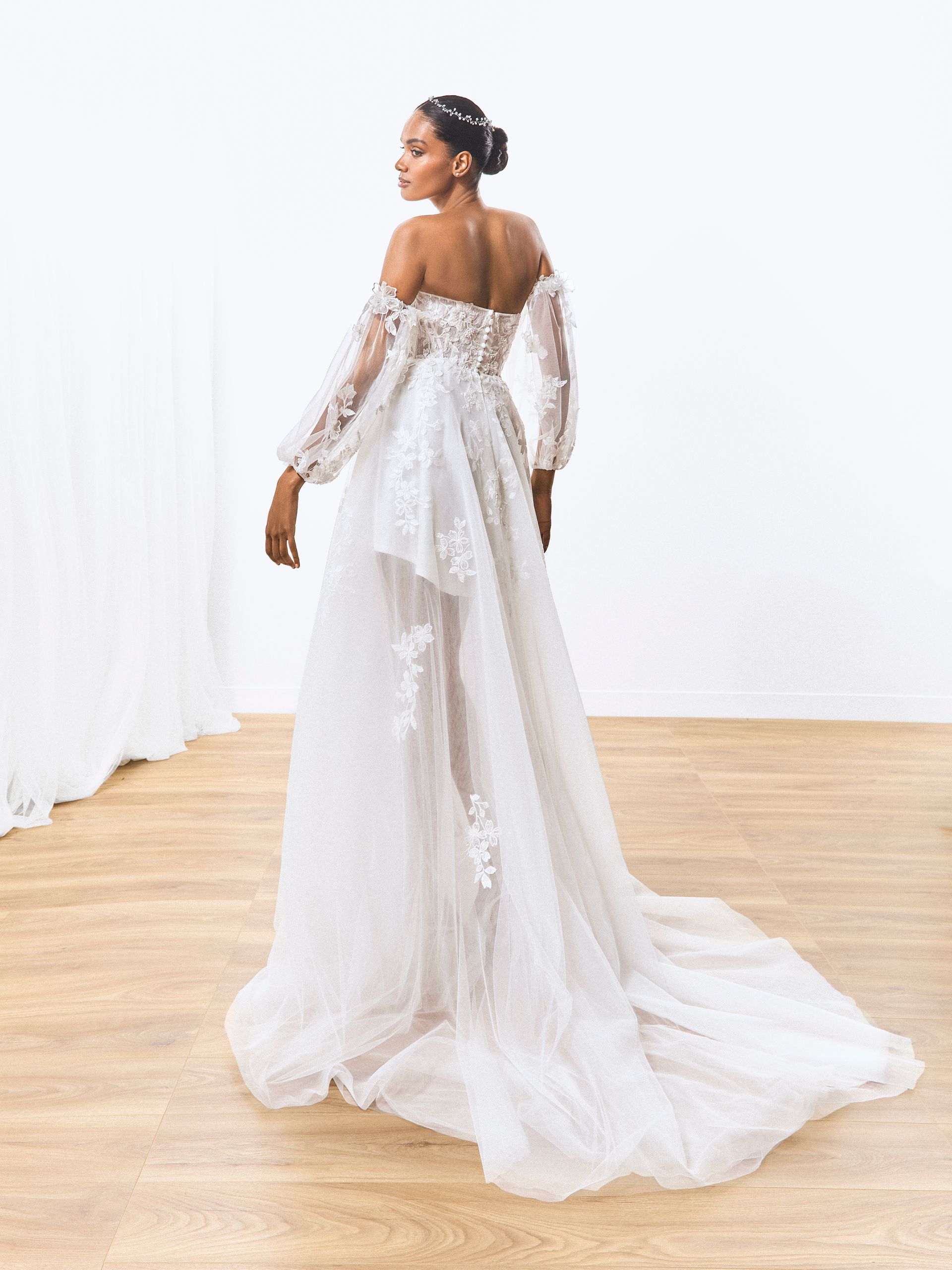 Donna con un abito da sposa bianco a spalle scoperte, di spalle, in una stanza luminosa con un lungo strascico.