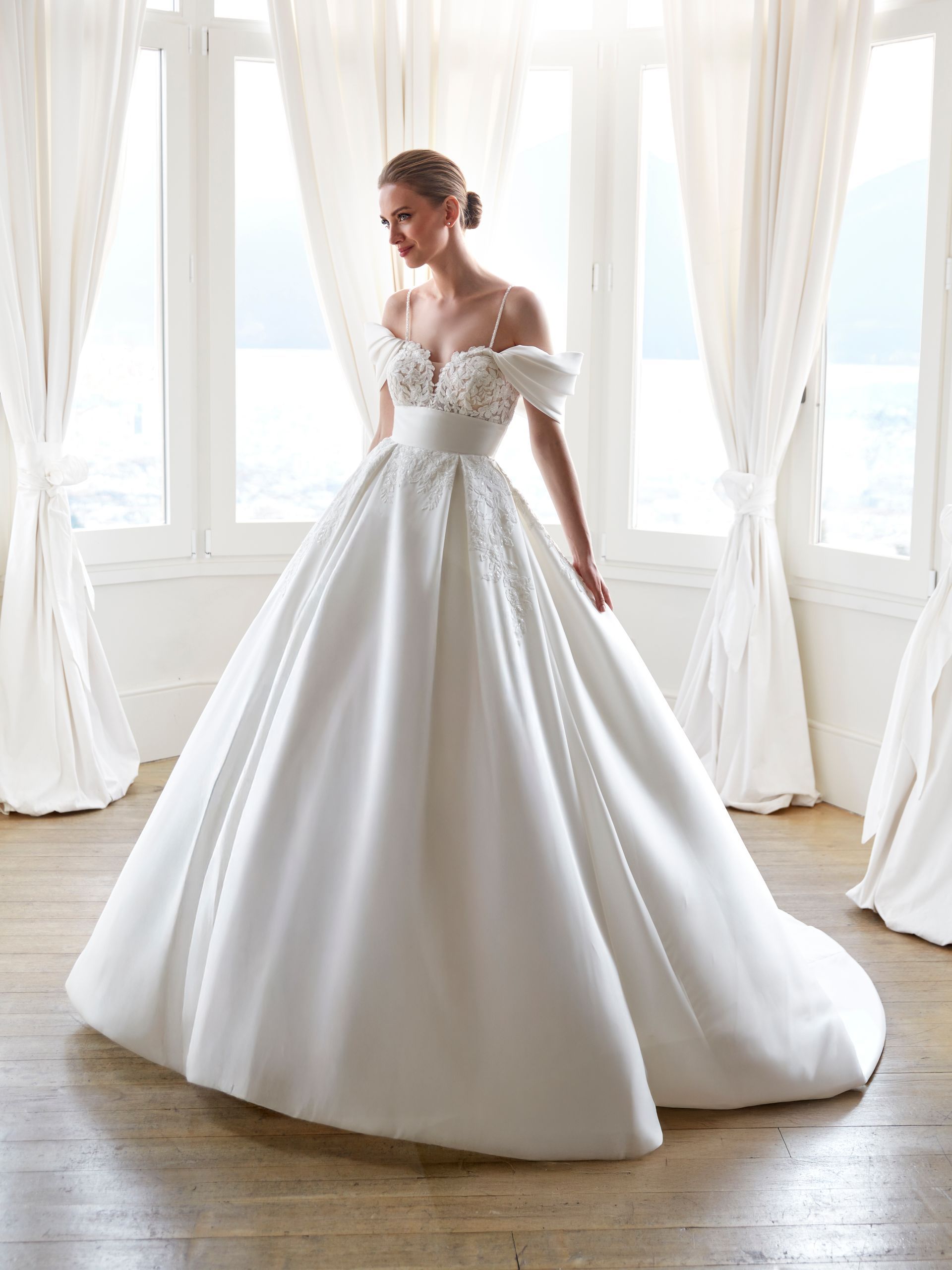 Sposa in abito da ballo in raso bianco, in posa vicino a una grande finestra con morbide tende bianche.