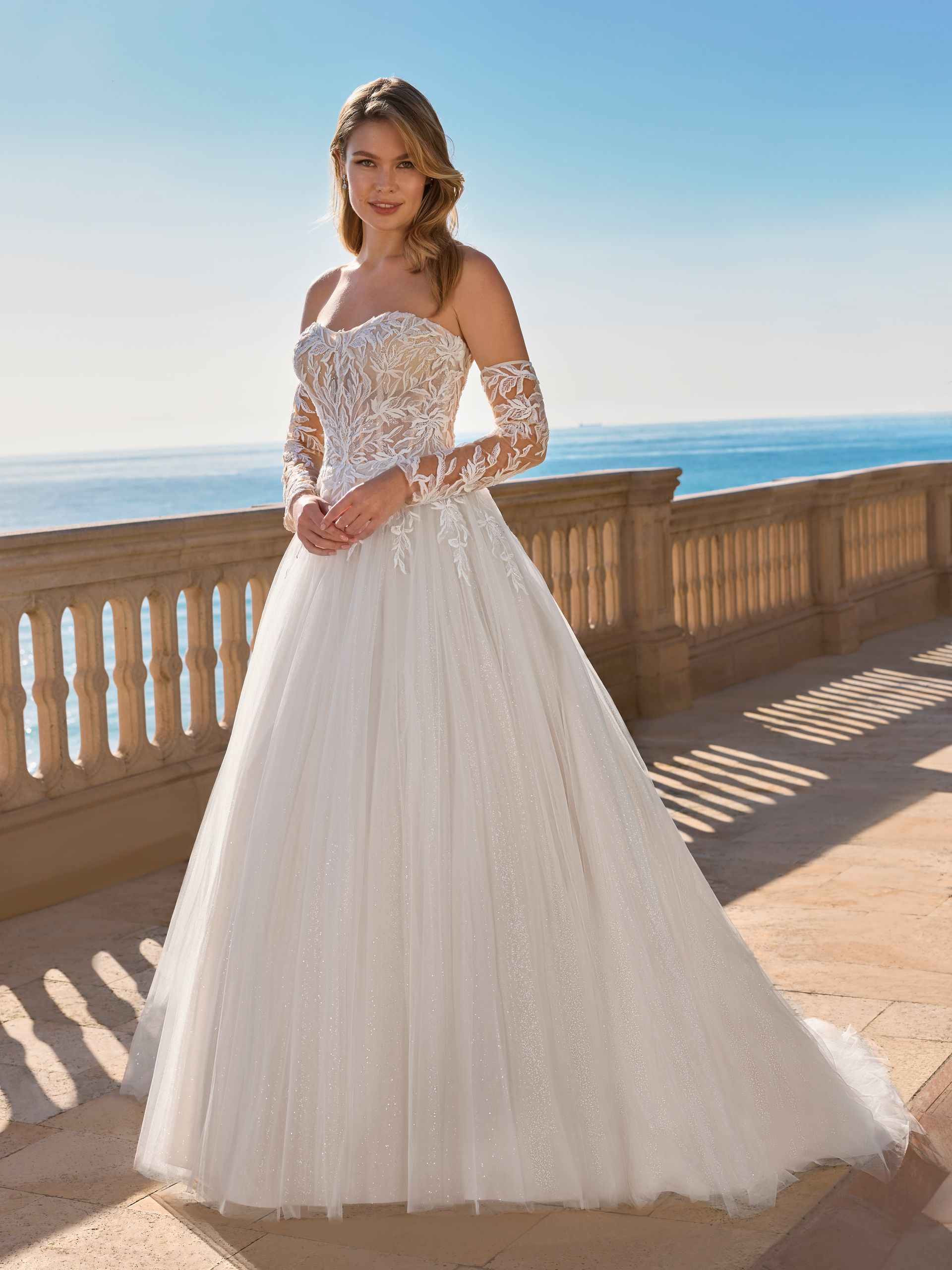 Donna in abito da sposa con maniche in pizzo, in posa su un balcone con vista sull'oceano.