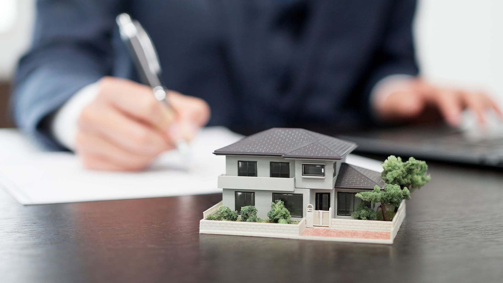 Miniature house on a desk with a person writing and using a laptop in the background.