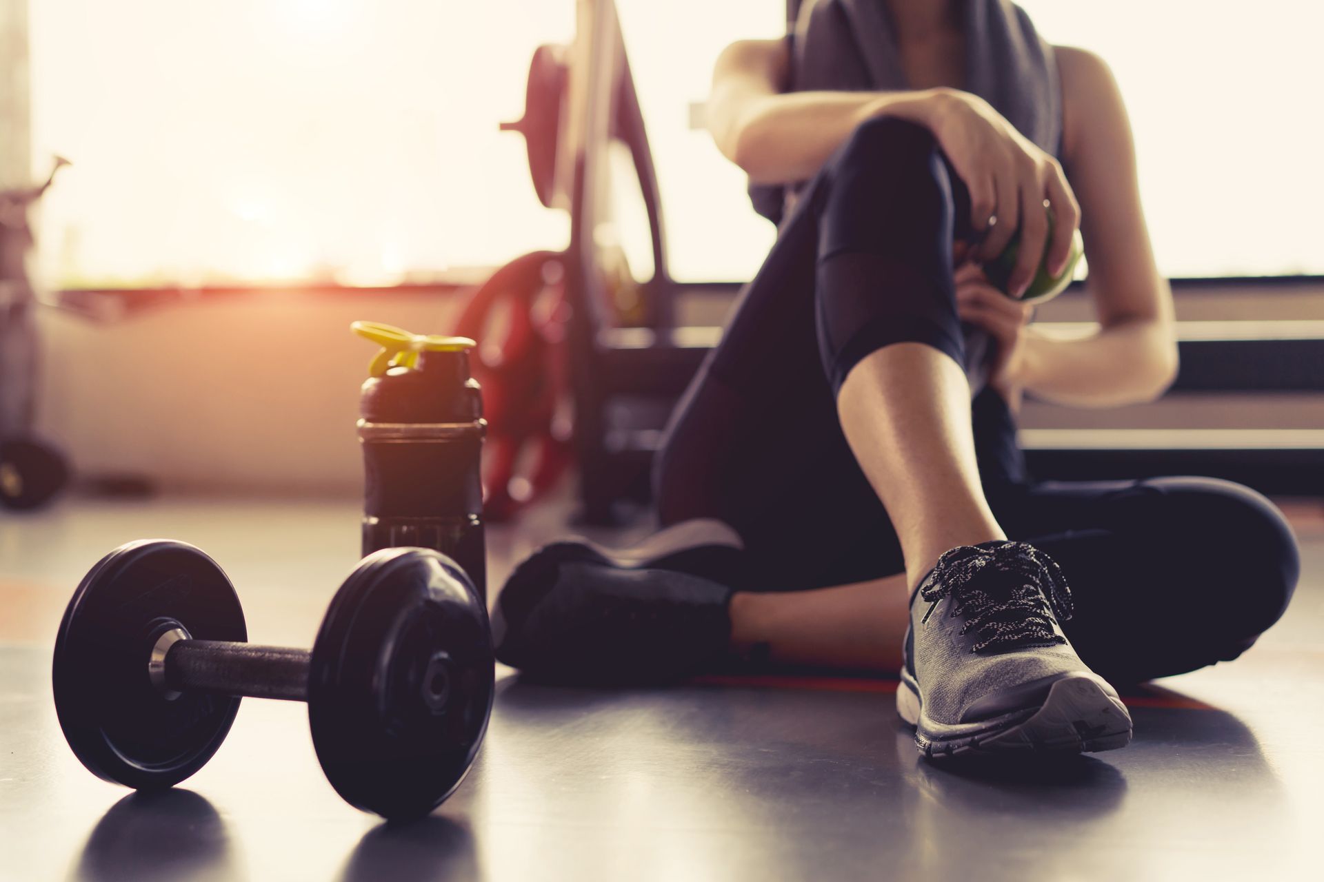 A woman is sitting on the floor in a gym next to dumbbells.