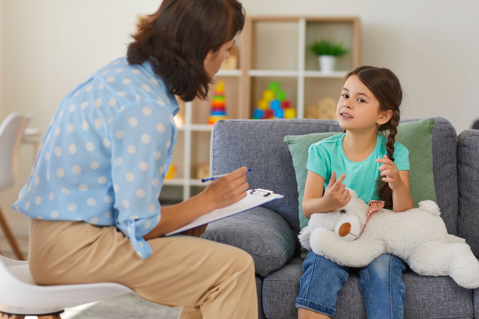 A woman is sitting on a couch talking to a little girl who is holding a teddy bear.