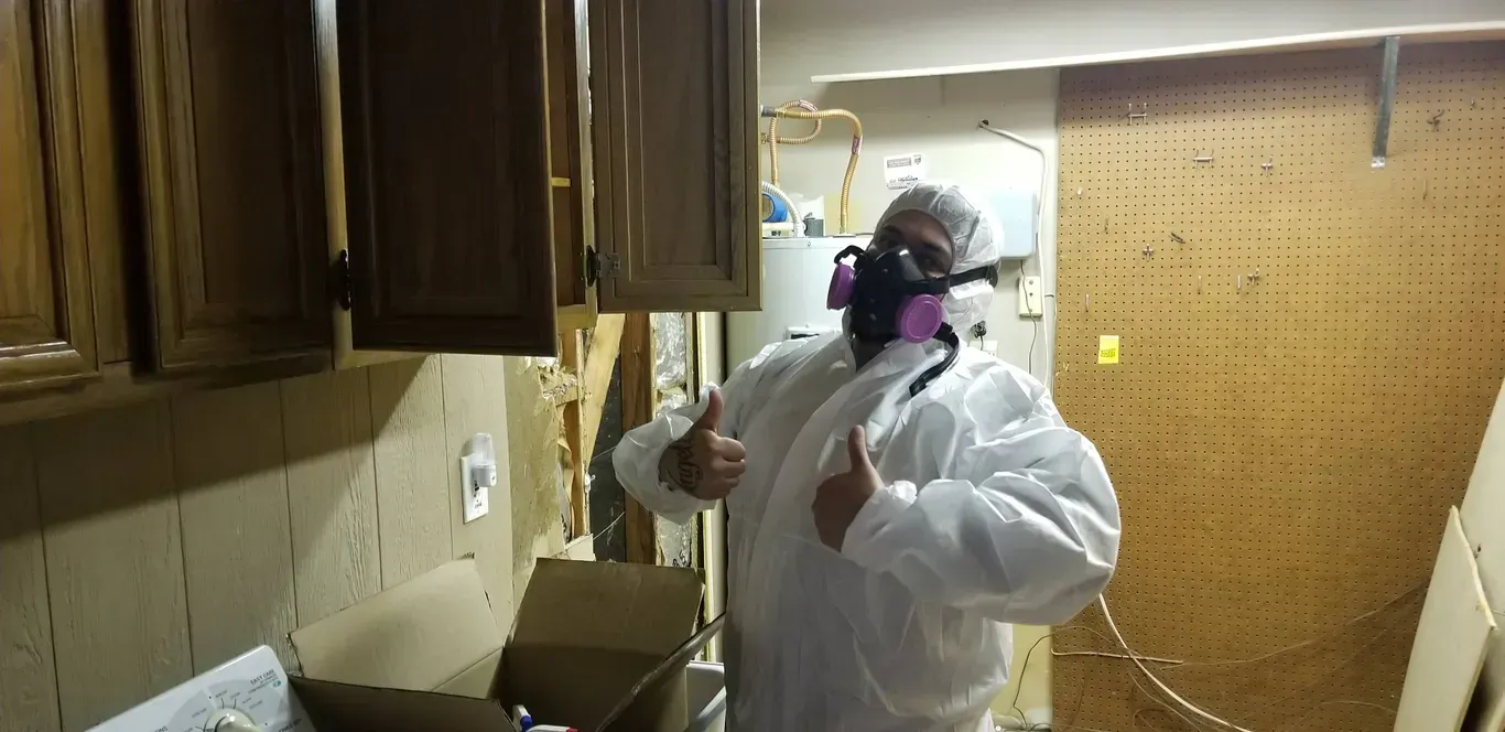 A man in a protective suit and mask is giving a thumbs up in a kitchen.